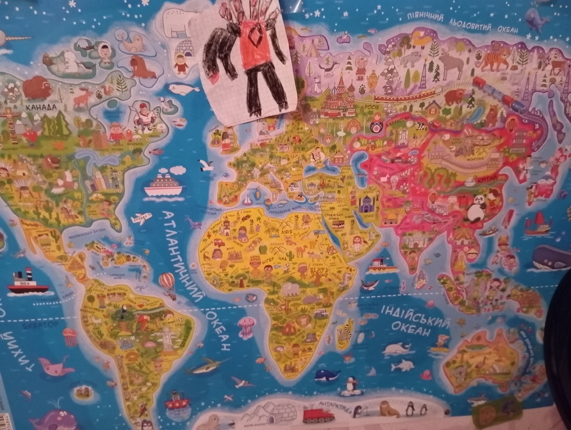 The image is a close-up, high-angle shot of a colorful world map poster, likely a children's educational map. The map is highly stylized and cartoonish, featuring illustrations of animals, landmarks, and people associated with different regions. It appears to be made of paper and is attached to a wall, as indicated by the slight curvature and the presence of a dark, out-of-focus object in the lower right corner, possibly a piece of furniture or an electronic device. A hand-drawn, two-dimensional figure, possibly a monster or a robot, is taped or affixed to the upper-middle section of the map, obscuring a portion of Eurasia. The figure is drawn in black marker with some red coloring. The map itself depicts continents and oceans with place names written in Cyrillic script. For example, "РОСІЯ" (Russia), "КАНАДА" (Canada), and "ІНДІЙСЬКИЙ ОКЕАН" (Indian Ocean) are visible. The overall impression is one of an informal, domestic setting, perhaps a child's room or a classroom. There is no identifiable context that specifically places this image in the city of Zaporizhzhia, Ukraine, beyond the use of Cyrillic script, which is common in Ukraine and other Eastern European countries. The image is well-lit, with the light source appearing to be from the top or upper-left.