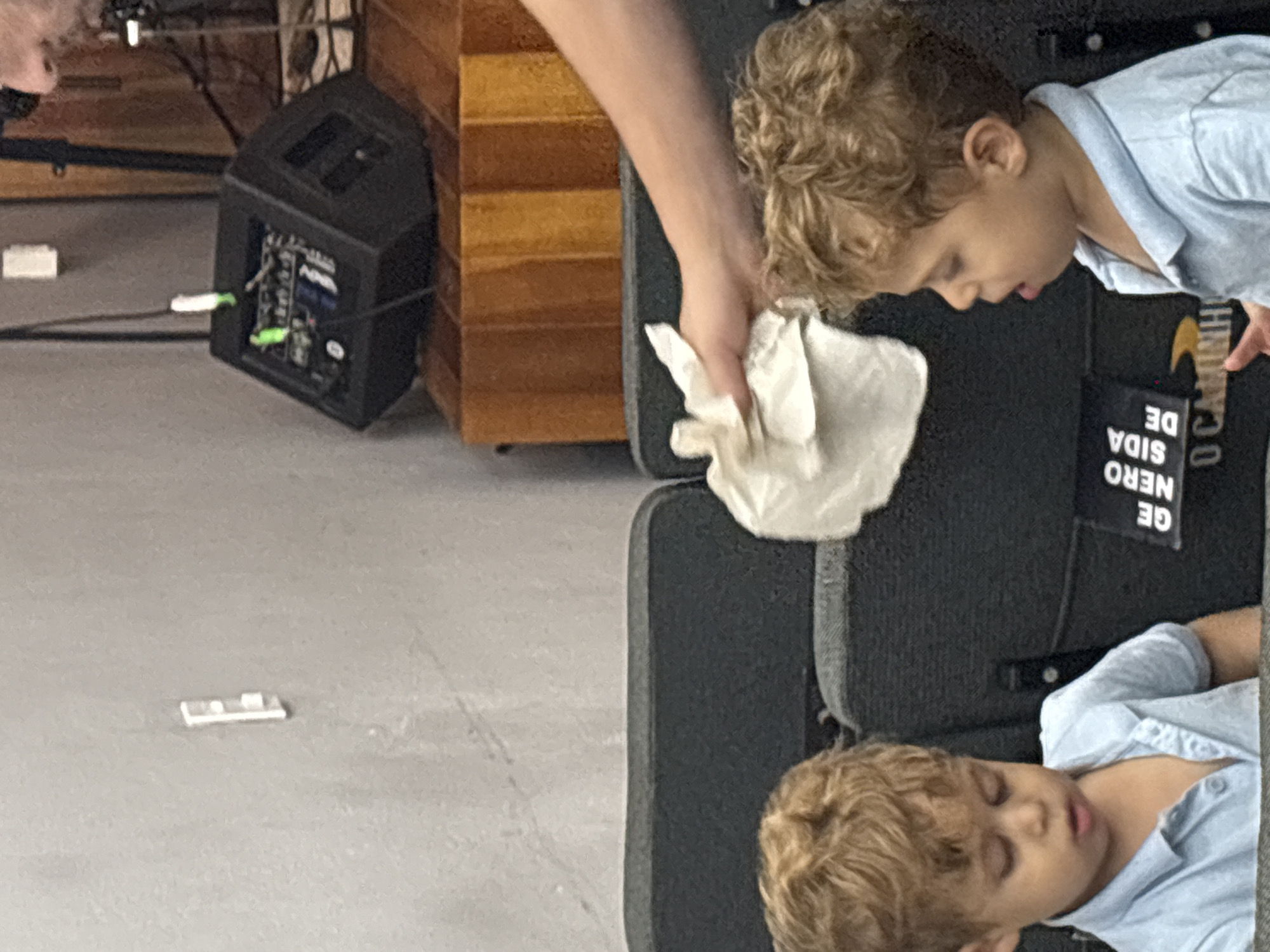 In São Paulo, Brazil, two young children with curly blonde hair are seated in dark blue theater seats. The child on the left wears a light blue collared shirt and looks down with closed eyes. The child on the right, also wearing a light blue collared shirt, looks to the side with their mouth open. A person's arm extends from the right side of the frame, holding a white tissue near the child on the right's head. Behind the children, a black speaker sits on a wooden surface. The wall behind the speaker is textured and gray. A black label with white text is partially visible on the seat between the children, reading "GE NERO SIDA DE O CAMINH".