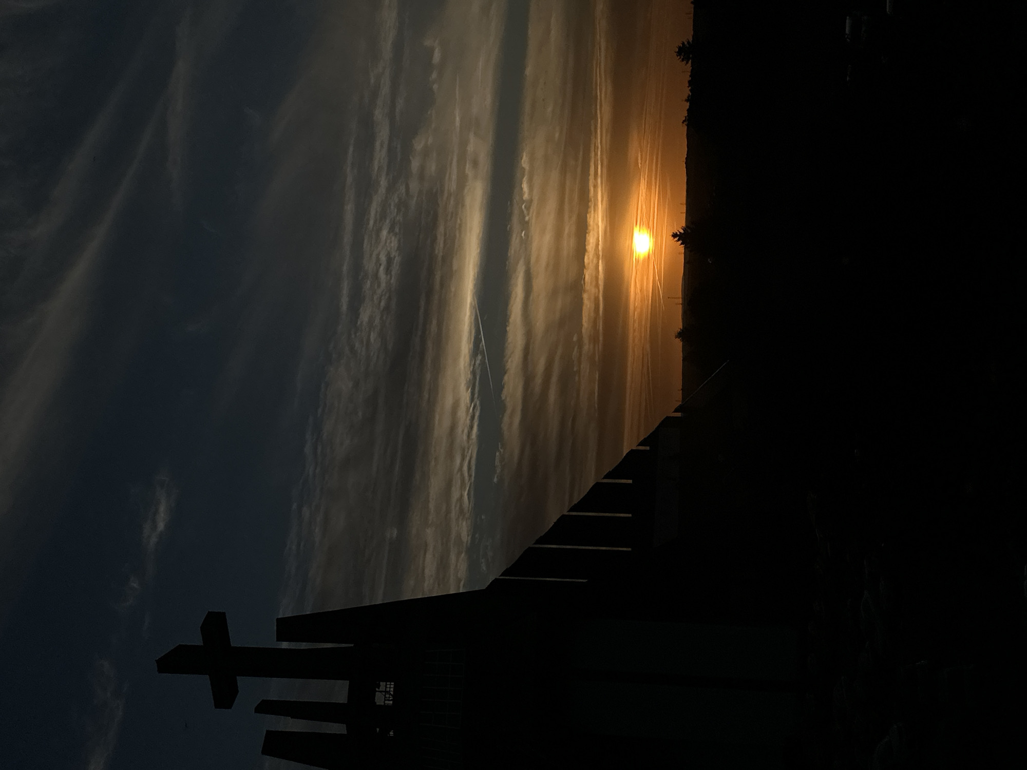 The image depicts a silhouette of a cross atop a modern architectural structure, possibly a church or monument, against a dramatic sunset sky. The structure is dark and angular, with vertical elements and a large cross positioned prominently. The sky is filled with streaks of clouds, illuminated by the setting sun, which appears as a bright, orange orb near the horizon. The lower portion of the image shows a dark, silhouetted landscape with sparse trees. Based on the architectural style and the presence of the cross, this scene is likely the Krzyż Milenijny (Millennium Cross) located in Świdnica, Poland. There are no people visible in the image. The overall atmosphere is solemn and evocative, capturing a moment at dusk.