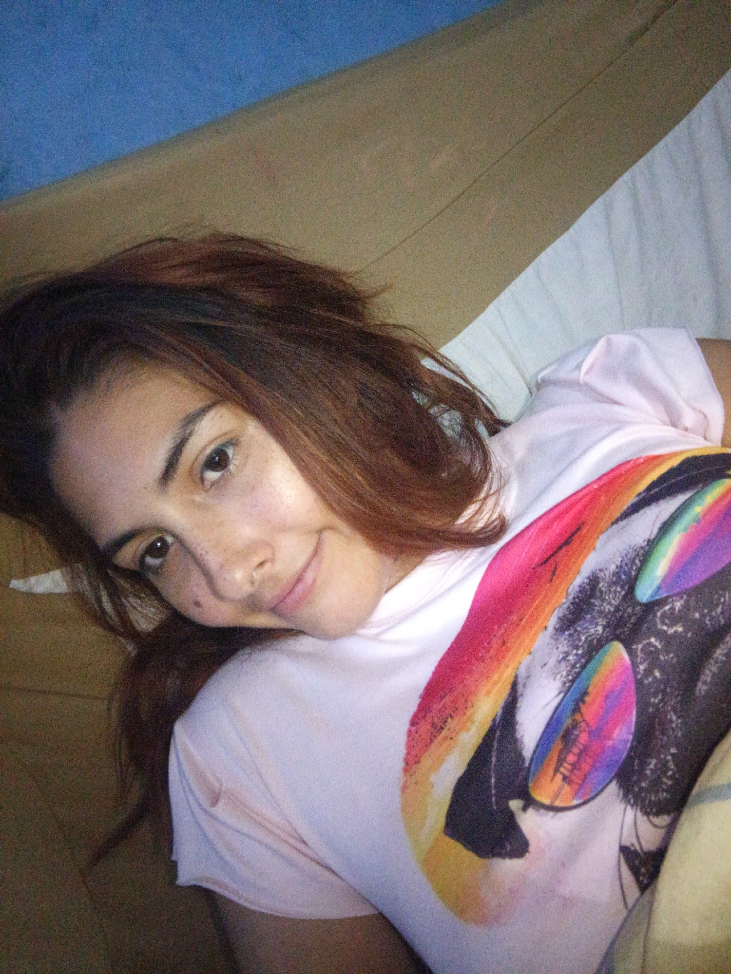 A young woman with long, reddish-brown hair is lying on a bed, looking at the camera. She is wearing a light pink t-shirt with a colorful, abstract graphic design on the front. Her head is tilted to the left, and she has a slight smile on her face. Her eyes are dark brown, and she has freckles scattered across her nose and cheeks. The background appears to be a bedroom with a blue wall and a tan headboard. A white sheet is visible beneath her. The image was taken in Juan Griego, Venezuela.