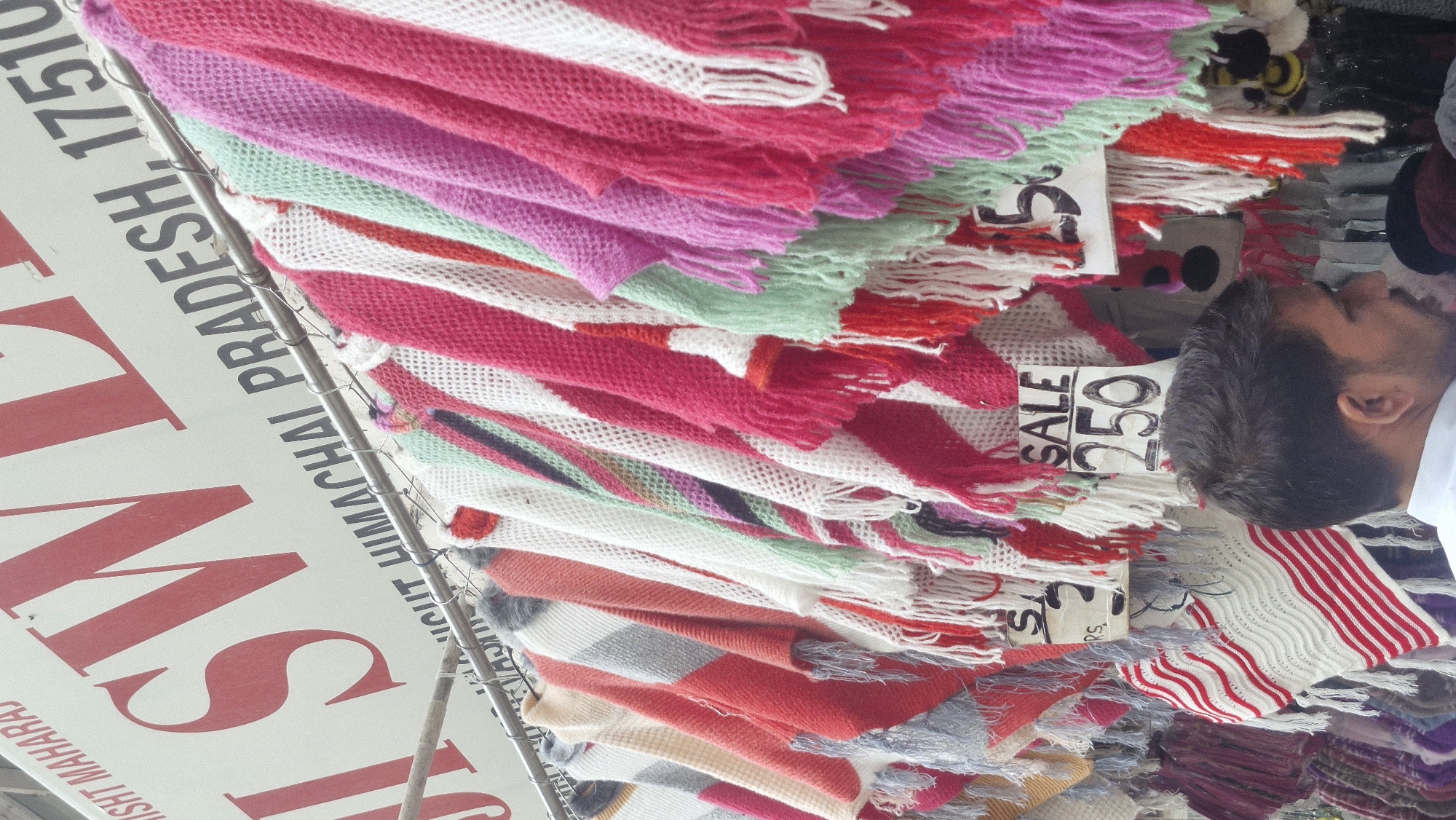 A street market stall in Vashisht, India, displays a vibrant collection of knitted shawls and ponchos in various colors, including pink, white, red, and green, with fringed edges. These items are hung on a metal rack, with some featuring price tags that read "SALE 250". A man, seen from the side, wearing a white shirt, is looking towards the merchandise. Behind the stall, a large sign with red lettering and "SW" prominently displayed is visible, along with text that appears to include "HIMACHAL PRADESH, 1751". The overall scene suggests a bustling marketplace.
