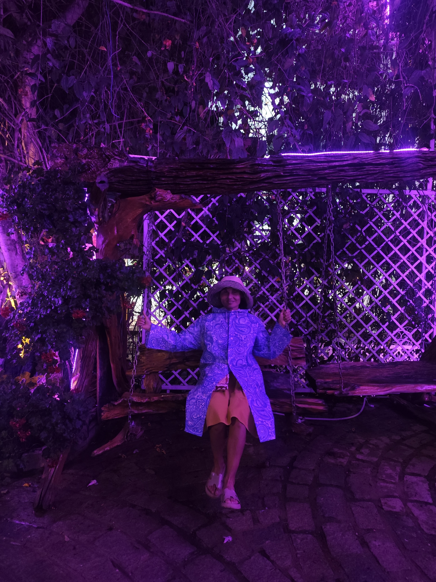 A woman sits on a wooden swing surrounded by purple lighting. The swing hangs from a wooden structure covered in vines and lattice. The woman is wearing a hat, a blue patterned coat, and orange shorts. The ground is paved with stone. The location is Da Lat, Vietnam.