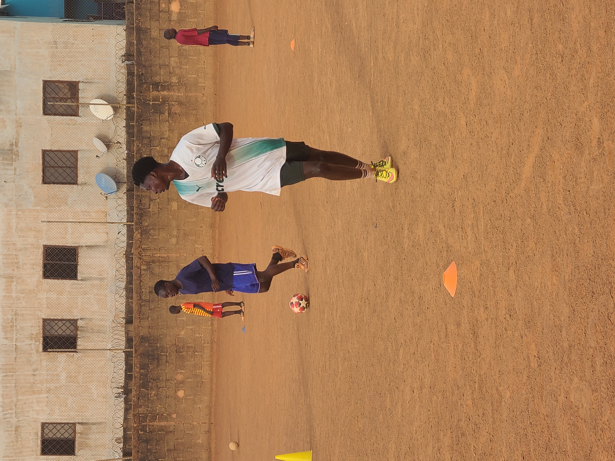 The image depicts a soccer field in Barakin Lerawi, Nigeria. Four individuals are present, presumably engaged in a soccer training session. One person is wearing a white and green jersey with dark shorts and yellow cleats, in what appears to be jogging action. Another individual is in a blue jersey and blue shorts, near a soccer ball. The other two individuals are further away, wearing red and orange, respectively. The field is dirt, with scattered cones, and surrounded by a wall. There are satellite dishes on the wall of the field. The background includes a building with windows.