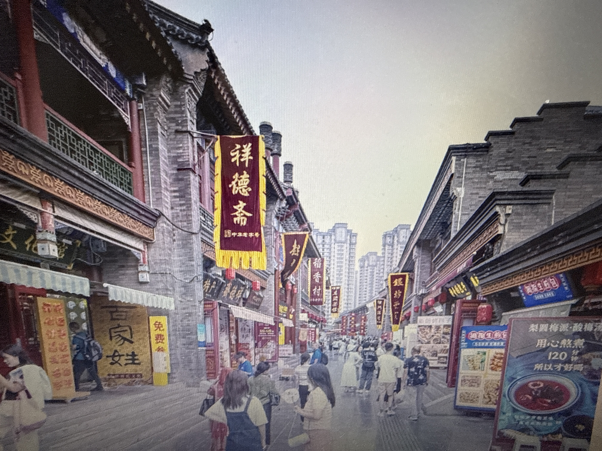 The image depicts a bustling street scene in what appears to be an East Asian city, characterized by traditional architecture and vibrant banners. The buildings lining the street have ornate roofs and brick facades. Several large banners with Chinese characters hang prominently, some in yellow and red, adding to the festive atmosphere. People are walking along the street, some are patrons of the shops, others are tourists. The street is paved and lined with shops displaying goods and signs with local language text. In the background, modern high-rise buildings suggest the coexistence of traditional and contemporary urban environments. The overall impression is one of a lively commercial area with a strong cultural identity. There is no indication of the location being Pushkino, Russia.