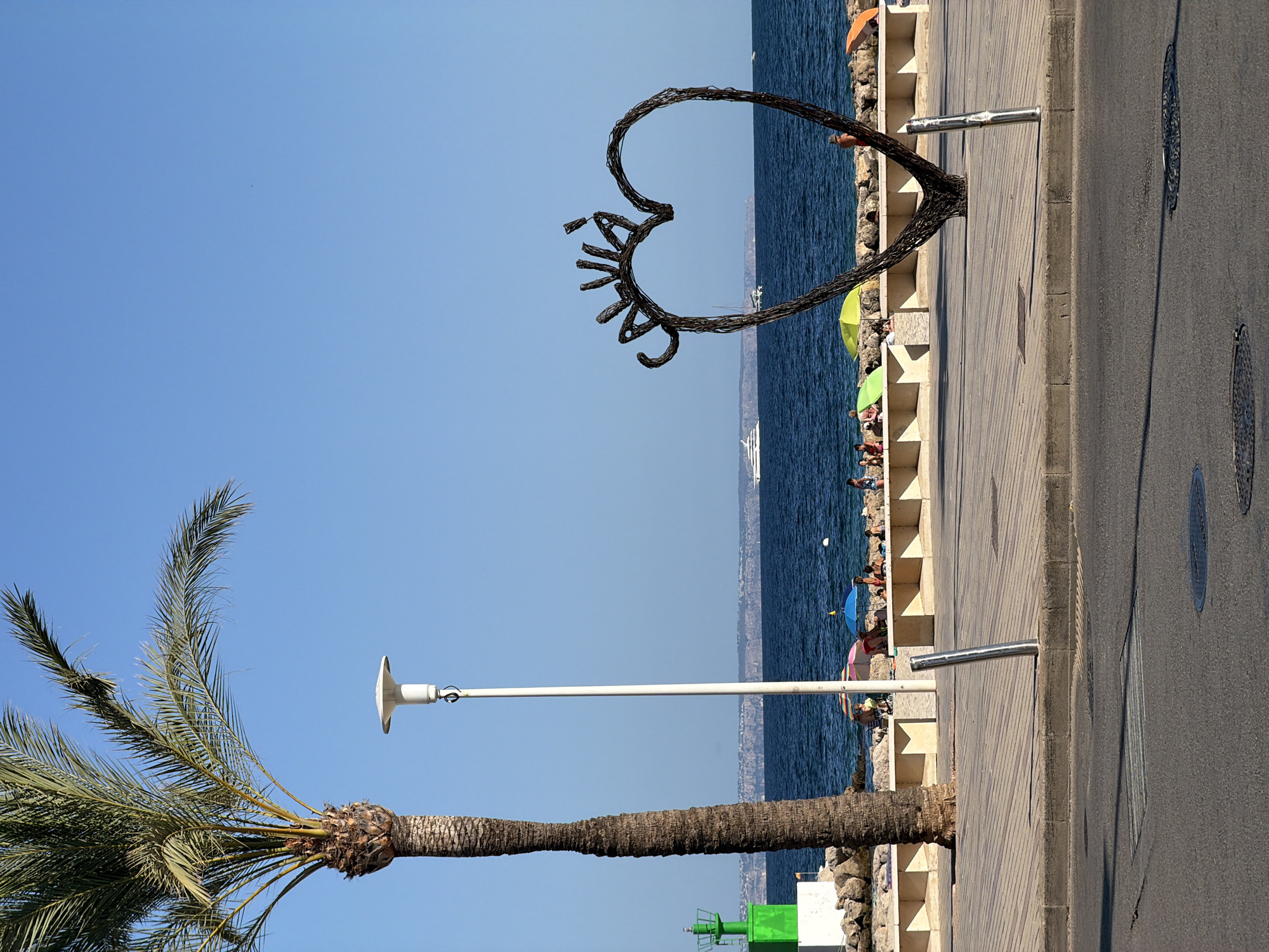 This image depicts a scene on a sunny day in Palmanova, Spain, likely along its promenade. A tall palm tree stands on the left, its fronds silhouetted against a clear blue sky. Beside it, a white lamppost with a modern, disc-shaped light fixture is visible. In the background, the deep blue Mediterranean Sea stretches to the horizon, with a distant coastline visible. Several boats, including a large white yacht, are scattered across the water.
On the sandy beach, people are enjoying the day under colorful umbrellas, some are sitting and relaxing, while others appear to be swimming or wading. A decorative, heart-shaped metal sculpture stands on the paved promenade, facing the sea. The sculpture is made of twisted metal and has the word "CALVIÀ" inscribed on its upper curve. The promenade itself is paved with light-colored tiles laid in a diagonal pattern and bordered by a concrete curb and a gray asphalt road. Several metal bollards are positioned along the promenade, and two manhole covers are visible on the road. To the left of the palm tree, a green and white lighthouse-like structure is partially visible. The overall atmosphere is relaxed and typical of a seaside resort.