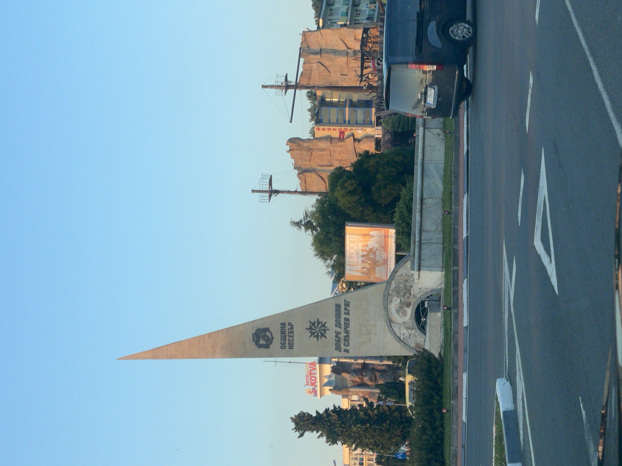 The image captures a street view in Nesebar, Bulgaria, dominated by a tall, triangular monument. The monument features inscriptions and emblems, including "ОБЩИНА НЕСЕБЪР" (Municipality of Nesebar) and "ДОБРЕ ДОШЛИ В СЛЪНЧЕВ БРЯГ" (Welcome to Sunny Beach). To the left of the monument, a statue of a person is partially visible. Behind the monument, a building with a facade resembling a rock formation and masts suggests a themed establishment, possibly a restaurant or entertainment venue, with the word "RESTAURANT" visible on its exterior. To the right of the monument, a billboard advertising "BEDROOM" with images of people is displayed. Further to the right, residential buildings are visible. In the foreground, a dark-colored van is parked on the right side of the road, and lane markings are present on the asphalt. The sky is clear and blue, indicating daylight.