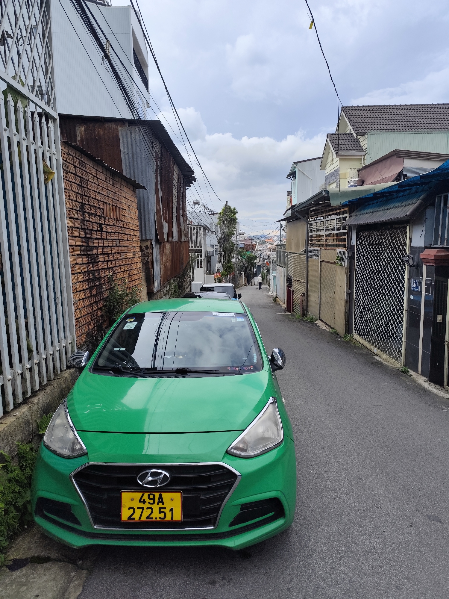 A green Hyundai i10 taxi with a license plate reading "49A 272.51" is parked on the right side of a narrow, paved street in Da Lat, Vietnam. The car is the primary subject in the foreground. The street is lined with buildings on both sides. To the left of the car is a white metal fence, a brick wall, and some overhanging power lines. The buildings on the right are a mix of residential and commercial structures, some with corrugated metal roofs and others with tiled roofs, displaying various architectural styles common in Da Lat. The sky is overcast with clouds. Further down the street, other buildings and distant hills are visible, indicative of the hilly terrain of Da Lat. A person is walking away from the camera further up the street, and another vehicle is parked on the opposite side of the street. The scene appears to be a typical urban street in Da Lat, characterized by its dense housing, narrow streets, and prevalent electrical wiring.