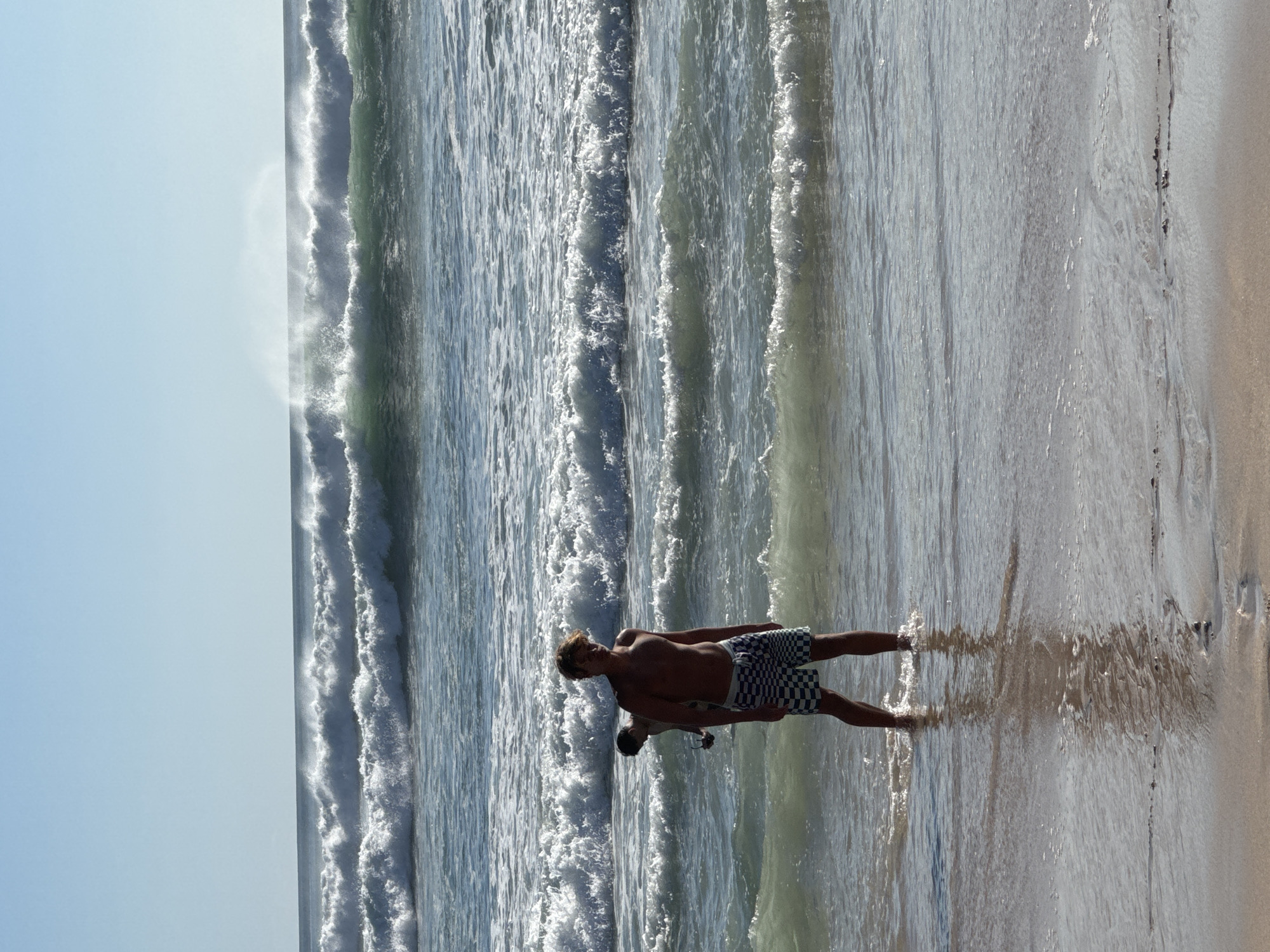 A young man is standing waist-deep in the ocean at Kuta Selatan, Indonesia. He is looking towards the camera and has a slight smile on his face. He is wearing checkered swim trunks. Behind him, another person is partially visible, appearing to be carrying a surfboard. The ocean is rough with large waves breaking in the background and smaller waves crashing around the man. The sky is clear and blue.