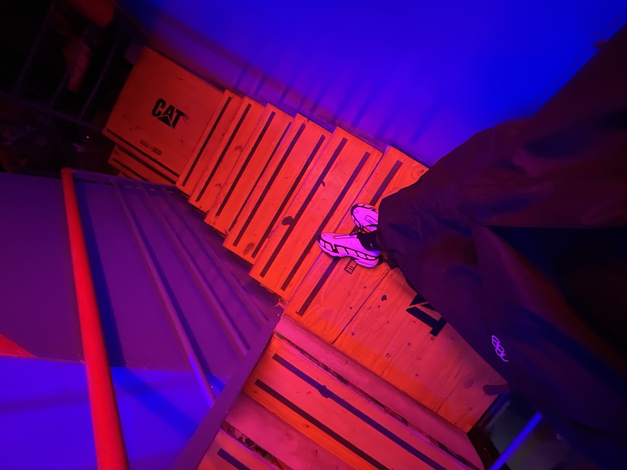 The image shows a view from above, looking down on a person wearing white and black athletic shoes, standing on what appears to be wooden steps or platforms. The steps are constructed from planks of wood with black banding, and one has the word "CAT" printed on it. The environment is illuminated with a mix of red, blue, and purple lighting, creating a dramatic and stylized effect. Some of the objects appear to be shipping crates or pallets. The location and city are not discernible from the image.