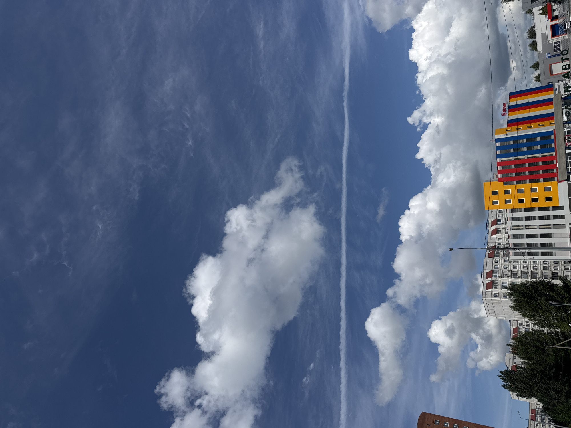 A wide shot captures a section of Tolyatti, Russia's skyline under a bright blue sky filled with dynamic clouds and a prominent contrail. In the foreground, several multi-story buildings line the bottom of the frame. The building on the far right is white with vertical grey accents and appears to be a commercial establishment with signage. To its left, a strikingly colorful building displays bands of yellow, red, blue, and white. Further left, a row of white residential buildings with red-trimmed roofs stretches across the mid-ground, partially obscured by dark green coniferous trees. A tall, slender street lamp stands to the right of the colorful building, and power lines crisscross the upper portion of the scene. The sky is a deep blue, populated by large, fluffy cumulus clouds and thinner, wispy cirrus clouds, with a distinct horizontal contrail drawn across the frame.
