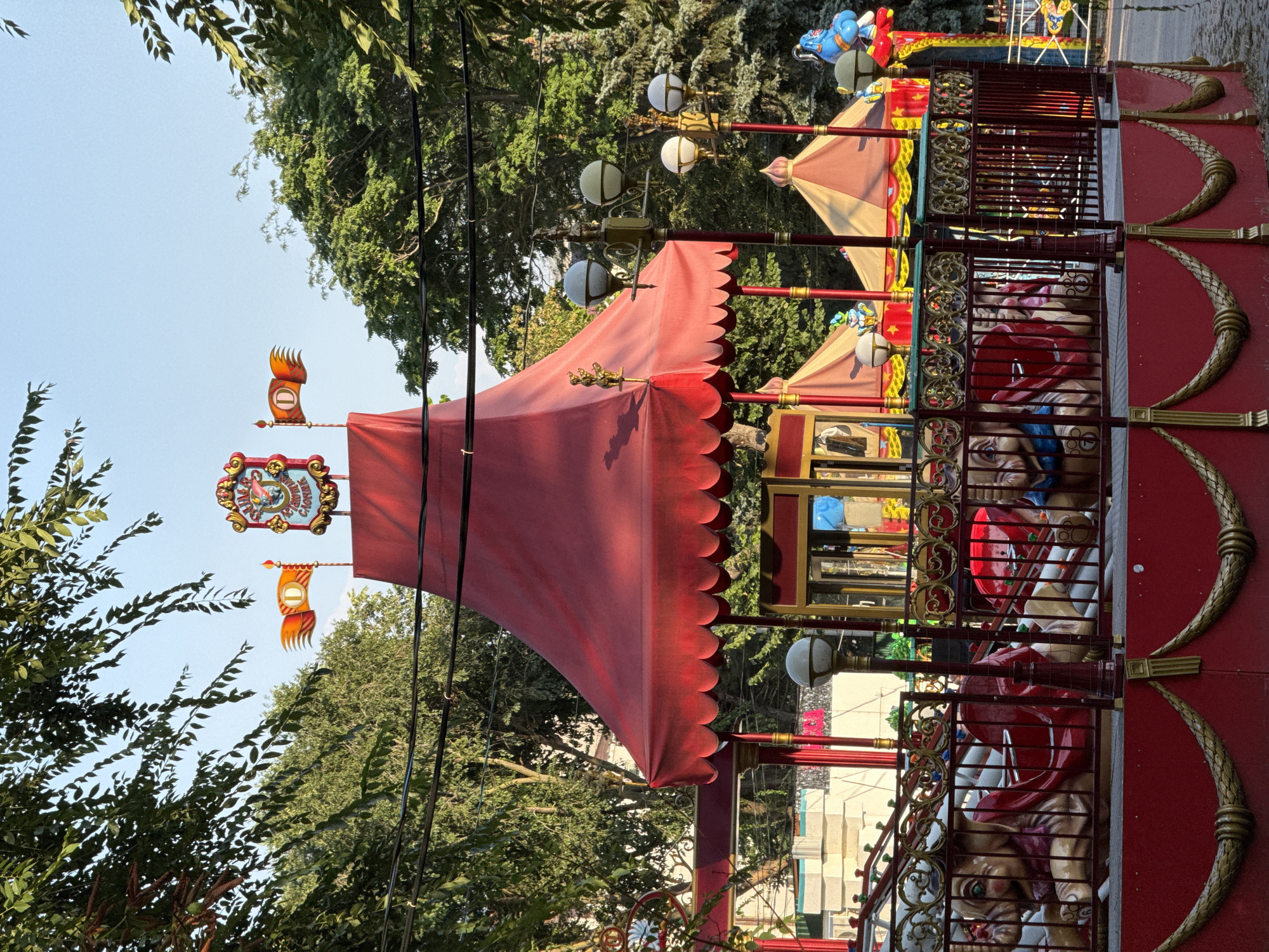 The image shows a close-up of a carousel in an amusement park, likely located in Rostov-on-Don, Russia. The carousel has a red canopy with scalloped edges, supported by red poles adorned with gold accents. The carousel animals are visible, appearing to be carved elephants with red saddles. A decorative gold railing surrounds the carousel platform. In the background, there are smaller tent-like structures and trees. The sky is clear and blue. The image captures a bright, sunny day.