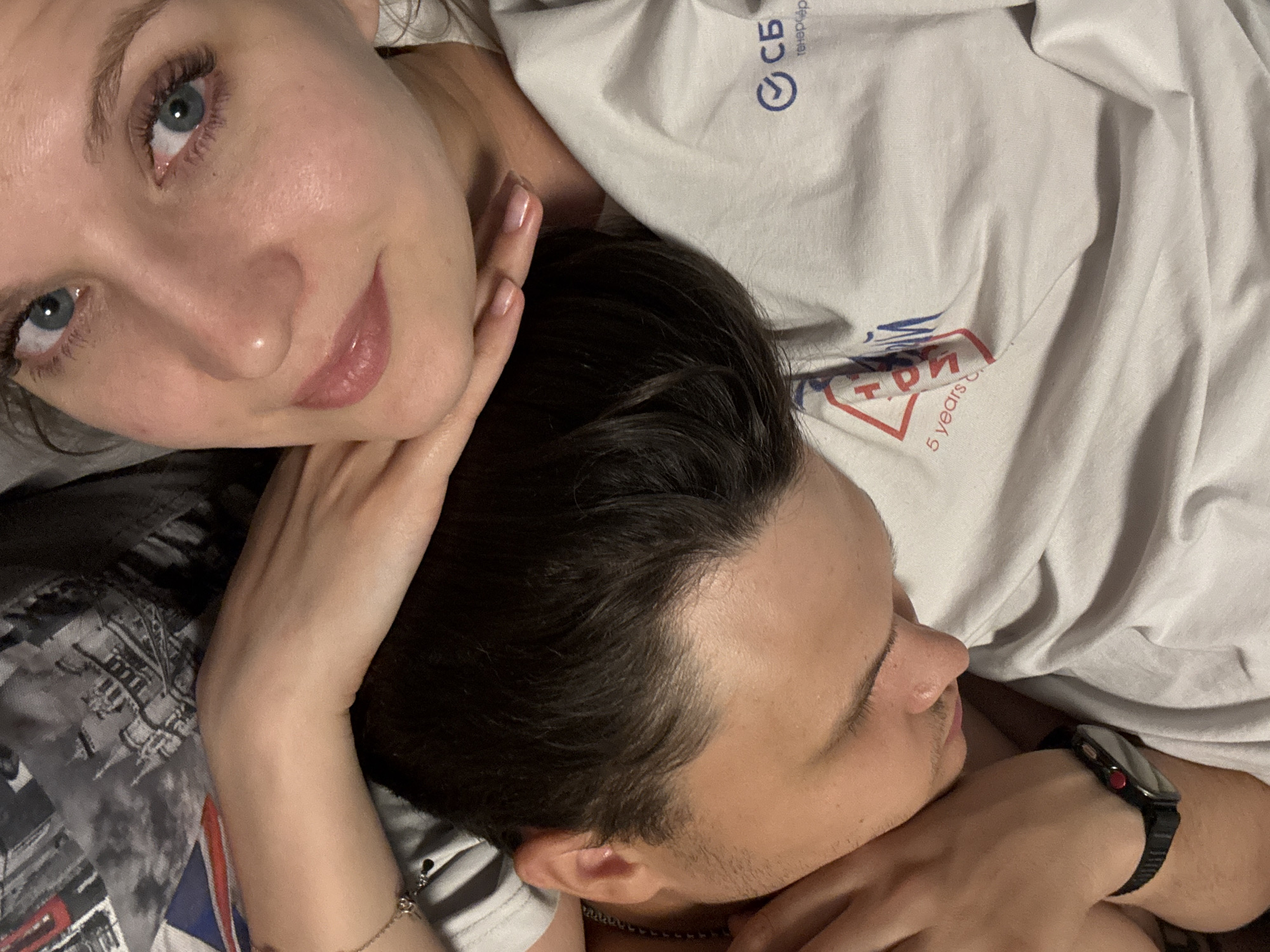 A young woman with blue eyes is lying close to a man with dark hair. The woman is looking directly at the camera with a slight smile. She has light skin and is wearing light pink lip gloss. Her hand is resting on the man's neck. The man has his eyes closed and is turned away from the camera. He is wearing a watch on his wrist. They are lying on what appears to be a bed with a white t-shirt visible. The t-shirt has some logos and text on it, including "СБ" and "5 years". It's not possible to confirm the location as Izhevsk, Russia, without further information.
