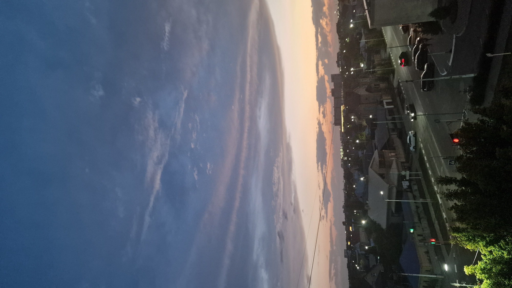 A high-angle, vertical shot captures an urban landscape at dusk. The sky is a gradient of deep blue to pale orange, with wispy clouds streaked with pink and white. Below, a city unfolds with houses, some featuring blue roofs, and taller buildings in the distance illuminated by lights. A paved road runs through the scene, with several cars, including a white sedan and a black SUV, driving on it. Streetlights are on, casting a glow on the road and sidewalks. Traffic signals are visible, with one displaying a red light. Lush green trees line the foreground, their leaves illuminated by artificial light. The overall scene is tranquil, with the transition from day to night evident.