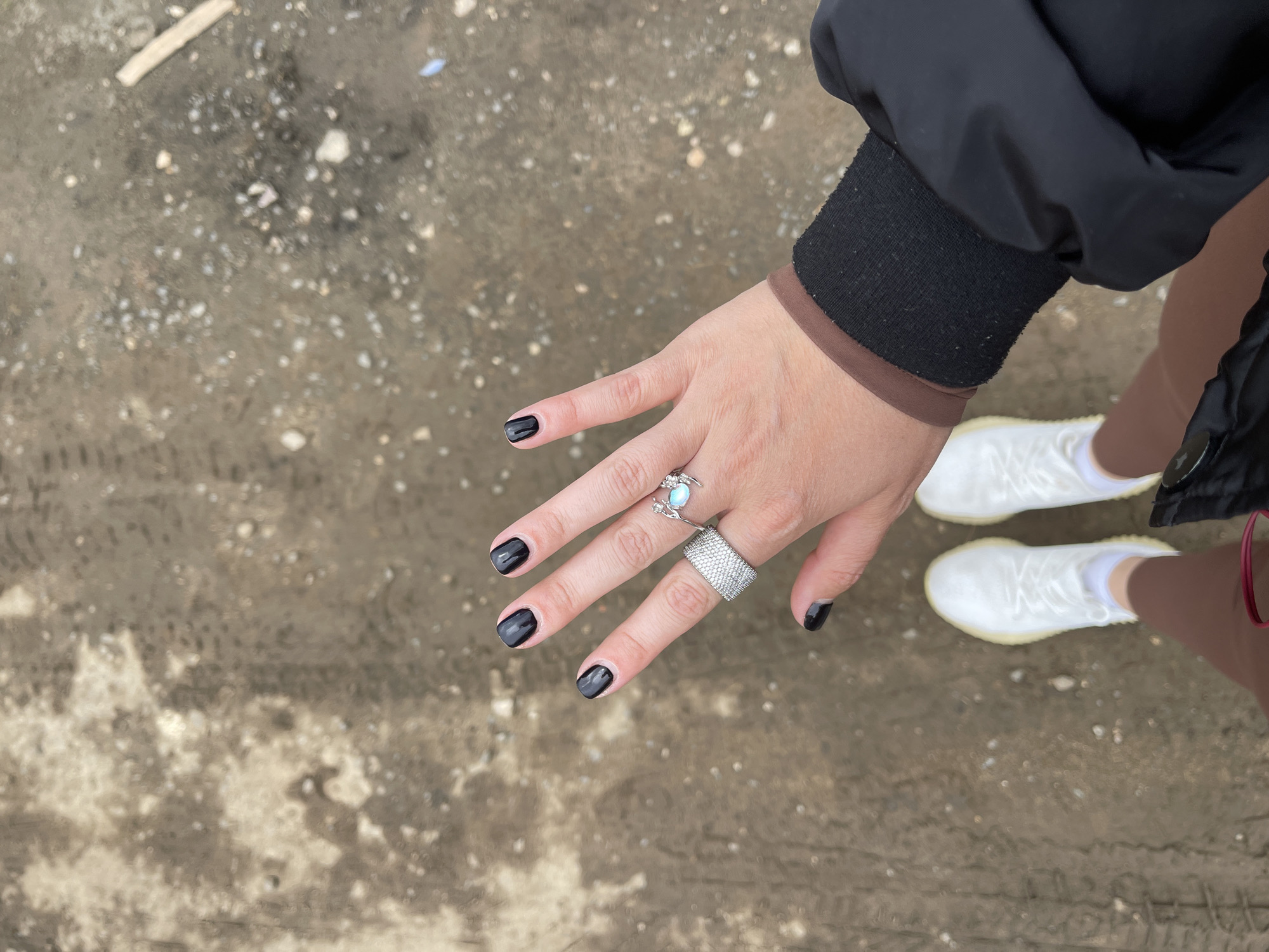 The image presents an aerial perspective, focusing on a person's left hand and feet positioned against a backdrop of dirt ground. The ground displays an uneven texture with apparent tire tracks.
The hand features short, polished fingernails with dark nail polish. The hand is adorned with two rings, one with a light blue stone on the middle finger and a larger, heavily textured ring on the ring finger. The person wears a dark jacket with a lighter brown inner lining at the sleeve and brown leggings. The feet are clad in white sneakers with white socks.
The location is Misaylovo, Russia.