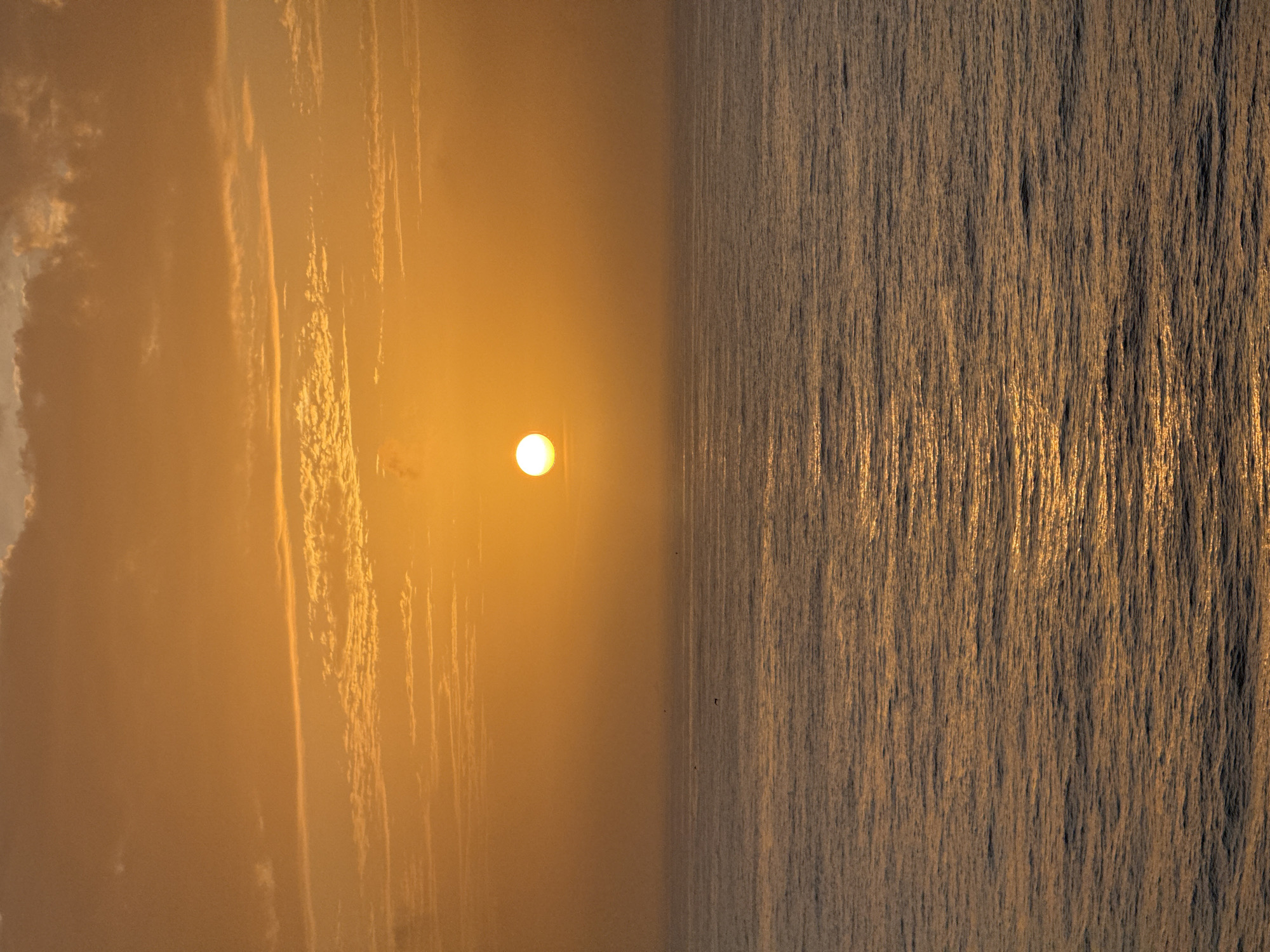 A landscape oriented photograph depicts a sunset over the ocean. The image is split into two distinct sections. The left half shows a pale orange sky with diffused clouds and the sun positioned slightly off-center. The right half consists of a textured expanse of water, reflecting the golden light of the setting sun. There are no visible people or structures. The location and city are unknown.
