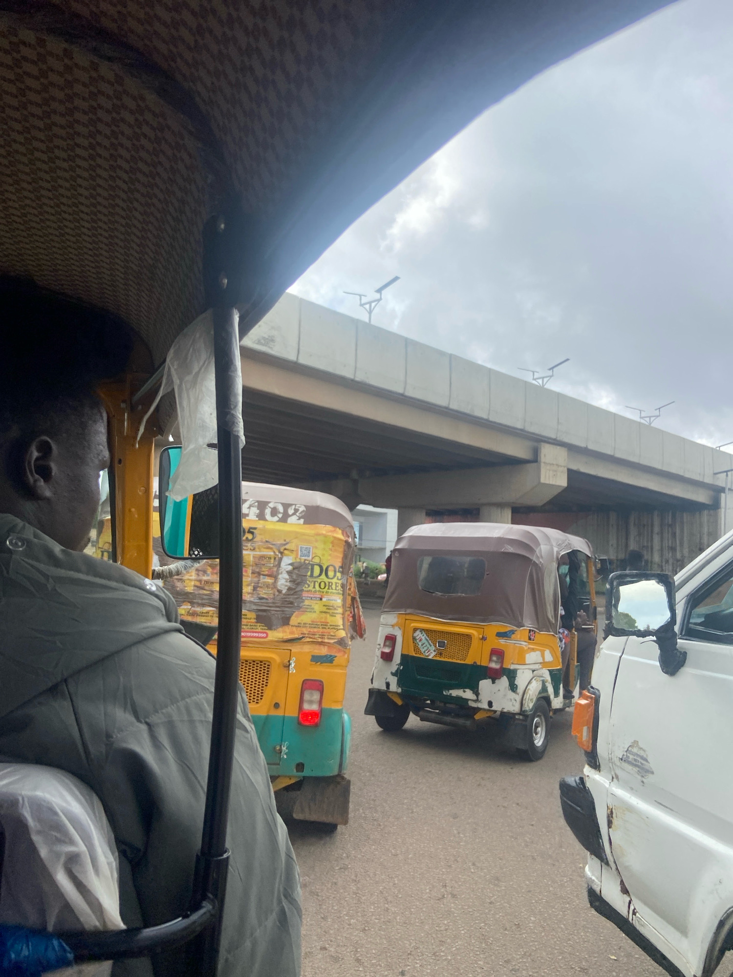 An over-the-shoulder view from inside a rickshaw in Jos, Nigeria, captures a traffic scene on a cloudy day. The rickshaw's tan and brown checkered roof is visible at the top of the frame. In front, the back of another yellow and green rickshaw is seen, decorated with various advertisements, one reading "DOS STORES". To the right, a white vehicle is partially visible, showing damage to its side and a side mirror. In the background, a concrete overpass looms, with streetlights mounted along its edge. The road appears to be asphalt, and the overall atmosphere suggests a busy urban environment.