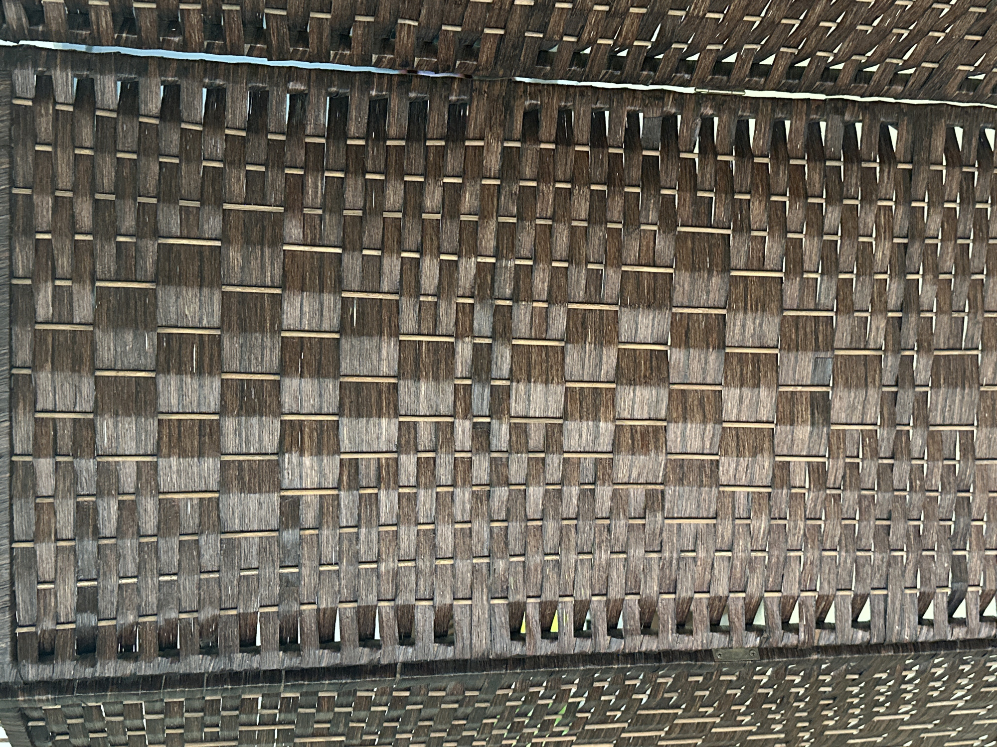 The image shows a close-up view of a dark brown woven screen, possibly made of rattan or bamboo. The screen is composed of horizontal and vertical strands, creating a textured, open-weave pattern. The lighting suggests it is daytime, and the background is slightly out of focus, showing hints of lighter colors that could be a wall or outdoor elements. No people or specific landmarks in Chalong, Thailand are visible.