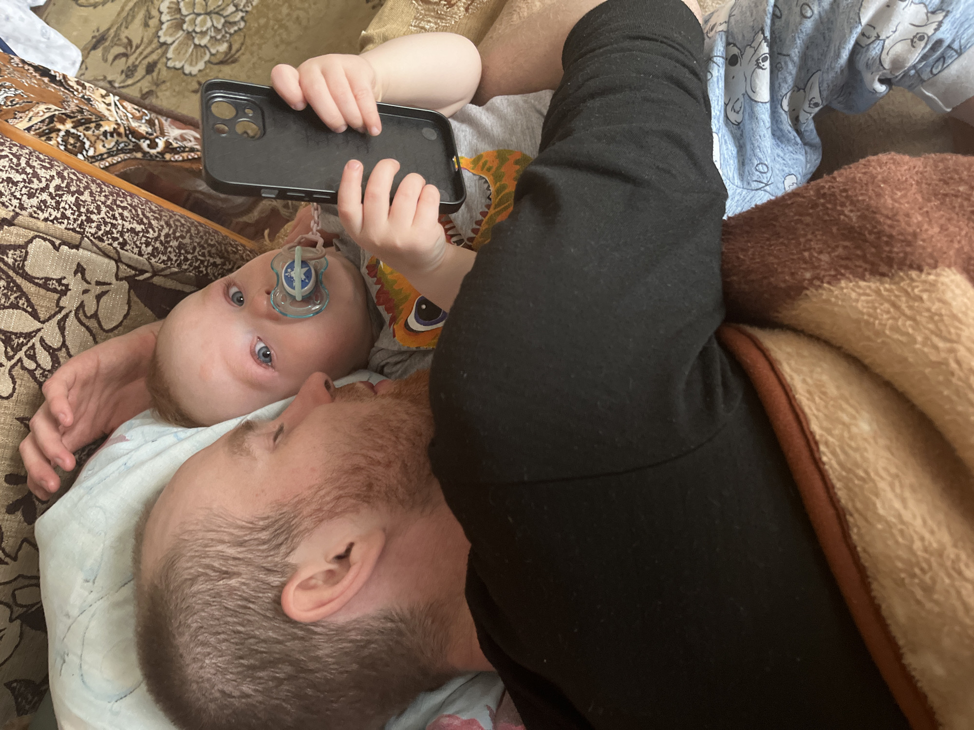 A top-down shot depicts a man lying on a patterned couch next to a baby, possibly in a domestic setting like a home in Сосново, Russia. The baby, with blue eyes and a pacifier, holds a black smartphone in its hands. The man is lying on a pillow with a light blue patterned cloth, and a brown blanket is partially visible. He appears to be interacting with the baby. The image suggests a casual, intimate moment between father and child.