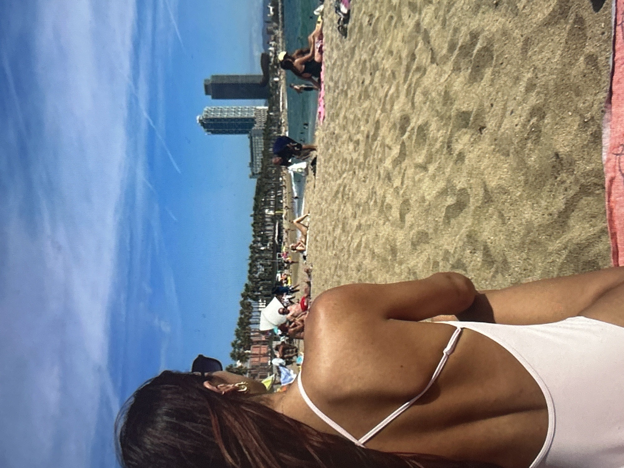 This media file captures a vibrant beach scene on a sunny day, viewed from a perspective on the sand. In the foreground, the back of a person is visible, looking out towards the ocean and city. This individual has long, dark hair, sunglasses perched on their head, and a gold hoop earring. They are wearing a light-colored, possibly white or pale pink, one-piece swimsuit with thin straps revealing a tanned back.
The midground showcases a busy sandy beach populated by numerous people. Many individuals are seen relaxing on towels, sitting, or standing near the water. A portion of a red or pink beach towel is visible on the right edge. Beyond the beach, the blue ocean contains more people swimming and enjoying the water.
In the background, a prominent cityscape rises along the coastline. Two distinct, tall skyscrapers stand out against the horizon, accompanied by other buildings and some vegetation, suggesting an urban coastal setting. The sky is a clear, bright blue with a few faint contrails or wispy clouds. The lighting indicates it is daytime, likely late morning or early afternoon, contributing to an atmosphere of leisure and summer activity. There is no visible text in the image.
