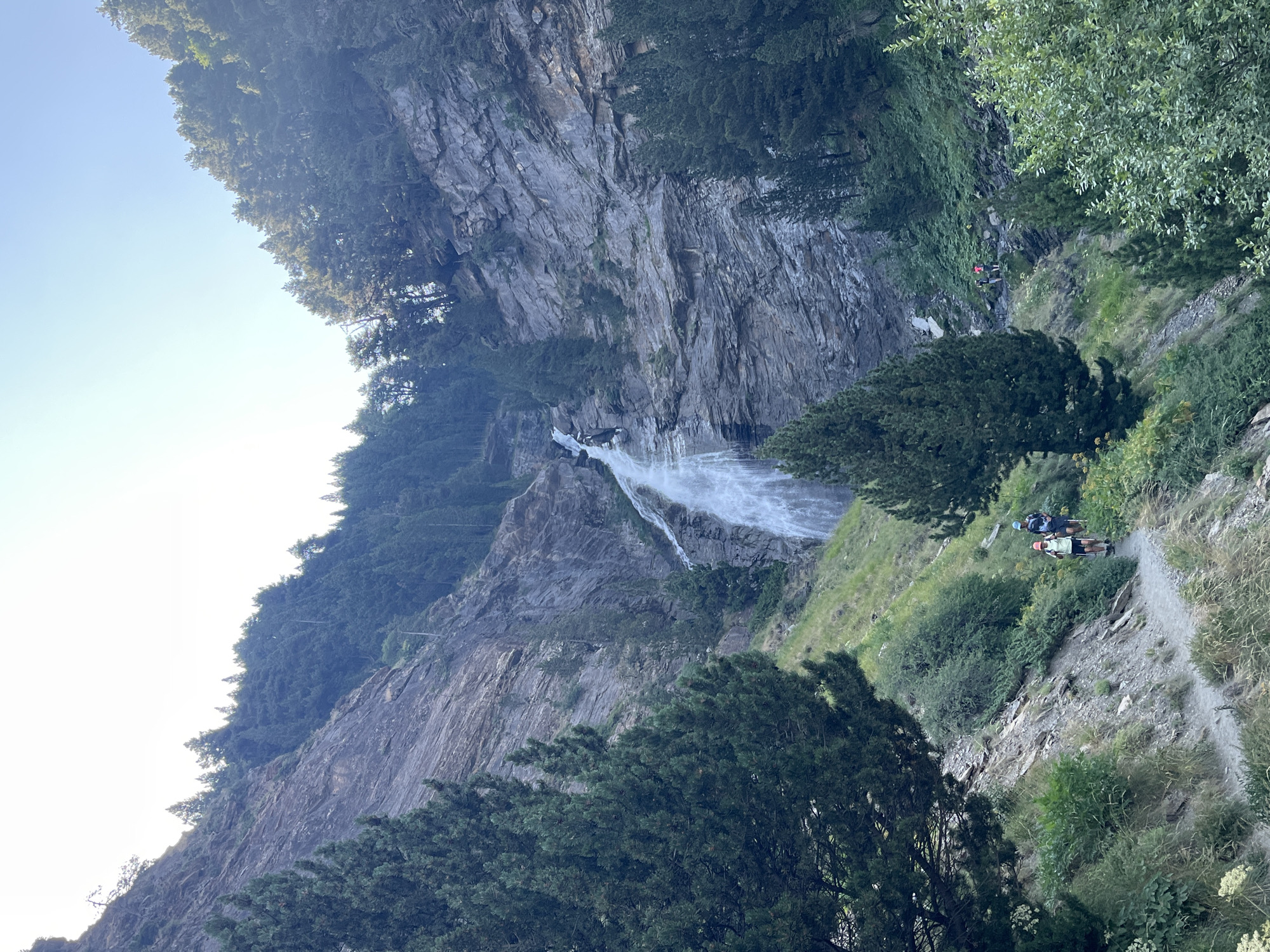 The image shows a tall, narrow waterfall cascading down a steep, rocky mountainside in the Pyrenees, likely near Benasque, Spain. Lush green pine trees and vegetation cling to the slopes. A well-worn dirt path winds through the lower portion of the scene, with two hikers, a man and a woman, walking along it. They are both wearing hiking attire and carrying backpacks, with the man using trekking poles. In the distance, further up the mountain and near the waterfall, two other figures are visible on the path. The sky is clear and bright blue, suggesting a sunny day.