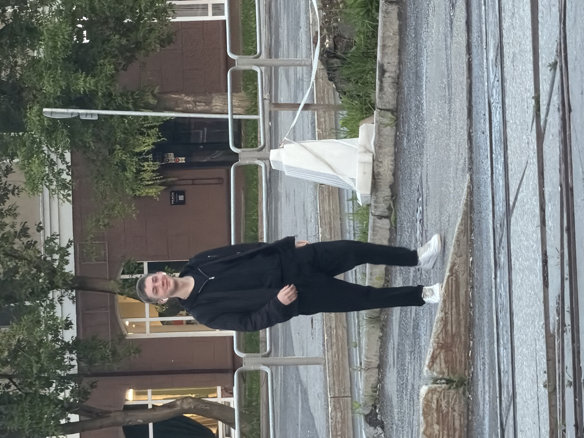 A young Caucasian male with short brown hair and fair skin is standing on a sidewalk in front of a brown brick building with large windows. He is wearing a black zip-up jacket over a black t-shirt and black pants, with white sneakers. He has white earbuds in his ears and is looking towards the camera with a slight smile. The building appears to be a commercial establishment, possibly a store, with a sign that reads "PRAGA" above a dark doorway. To the left of the building is a tree with green foliage, and to the right, another tree with similar foliage and a street sign. A metal fence runs along the edge of the sidewalk. In the foreground, there are tram tracks and a concrete curb. The environment suggests an urban setting, likely in Perm, Russia, based on the architectural style and signage.