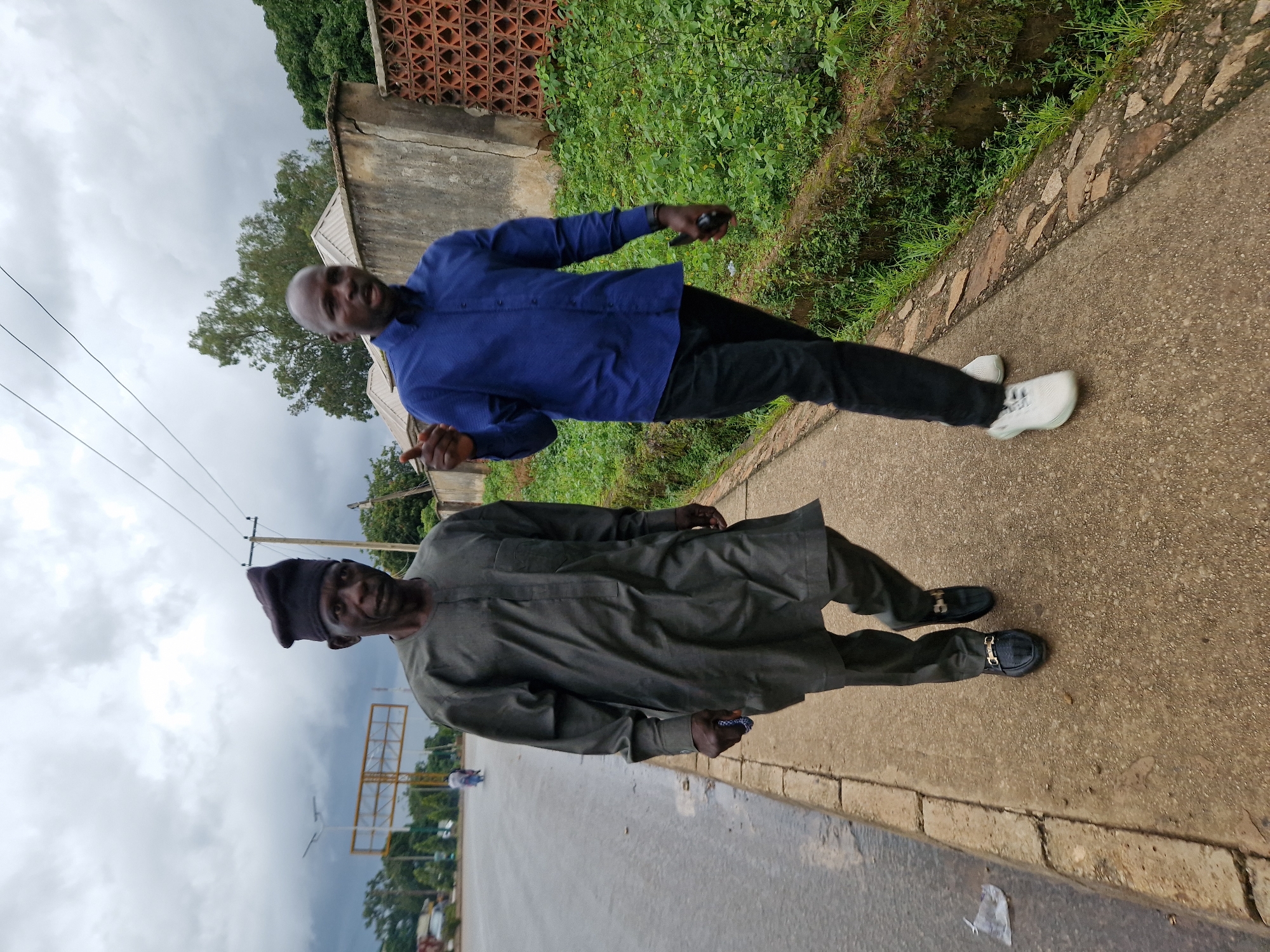 Two men are walking on a sidewalk in Jos, Nigeria. The man on the left is older and wearing traditional clothing, a dark grey tunic and pants with a matching cap. He has a stern expression on his face and is holding a small blue object in his right hand. The man on the right is younger and wearing a blue button-down shirt and black pants. He is gesturing with his right hand and appears to be speaking. The sidewalk is concrete and runs alongside a road. There is lush green vegetation on the right side of the sidewalk, bordering a wall. In the background, there are trees, a building, and a road with some traffic. The sky is overcast and appears to be cloudy.