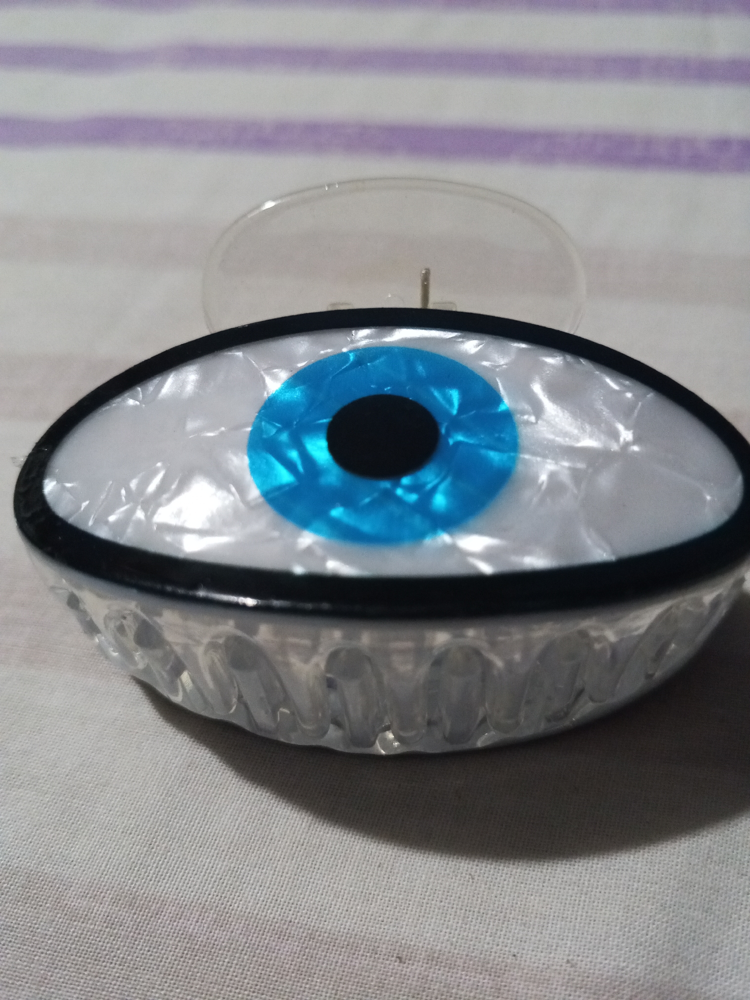 The image shows a hair claw clip shaped like an eye. The eye has a white, pearlescent background with a blue iris and a black pupil. The claw part of the clip is made of clear plastic. The clip is sitting on a surface with light purple and white stripes. The location is Unknown, Unknown.