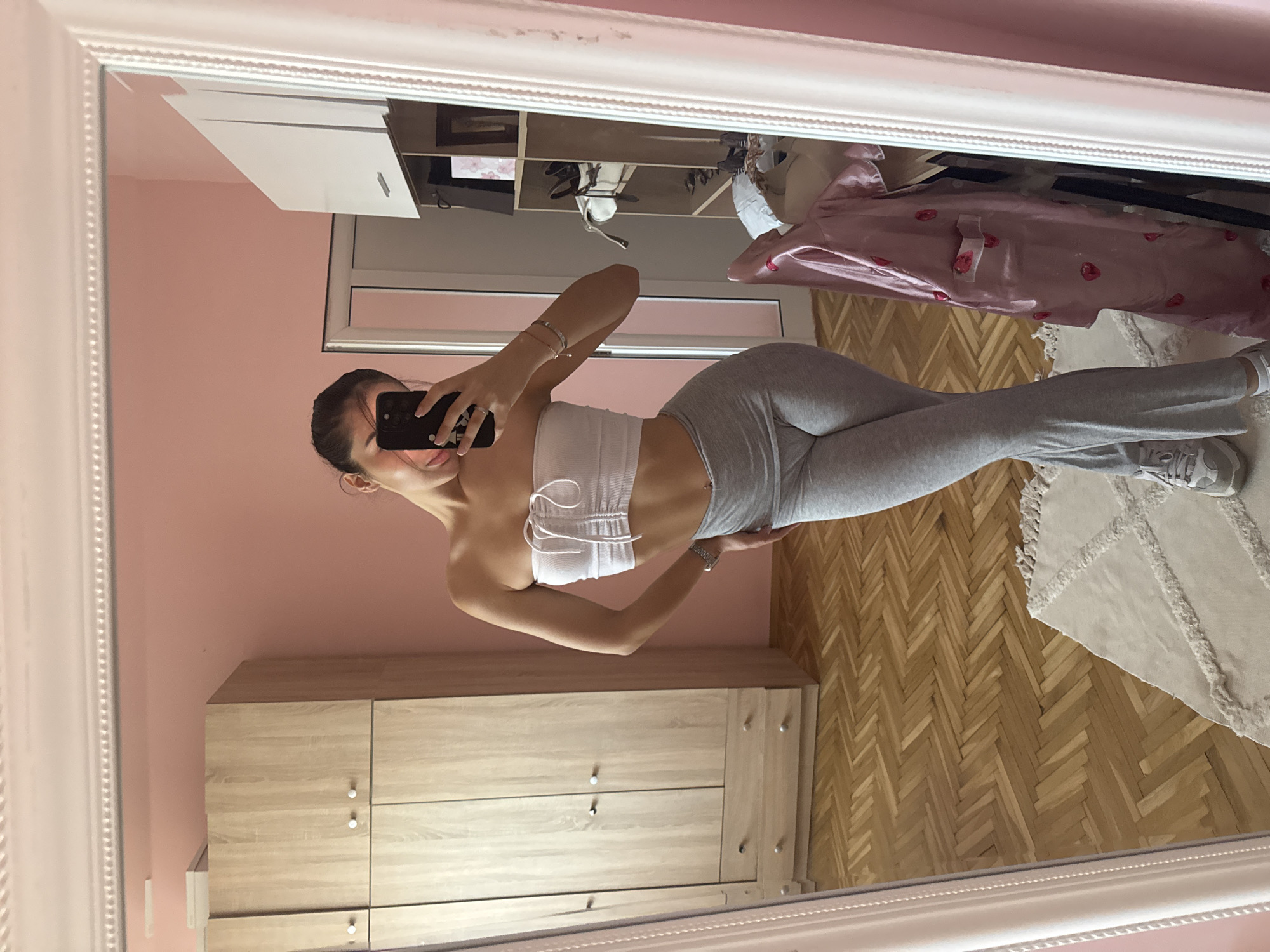 A young woman with dark hair pulled back is taking a mirror selfie in a bedroom. She is wearing a white strapless top and grey flared pants. She is standing in front of a wooden wardrobe and a pink wall. In the mirror, a shelf unit with various items, a pink satin robe with a strawberry pattern, and part of a white door are visible. The floor is made of light-colored wood laid in a herringbone pattern, and a cream-colored rug with a textured pattern is on the floor. The image appears to be taken indoors, likely in an apartment. There is no discernible indication of the location being in Plovdiv, Bulgaria.