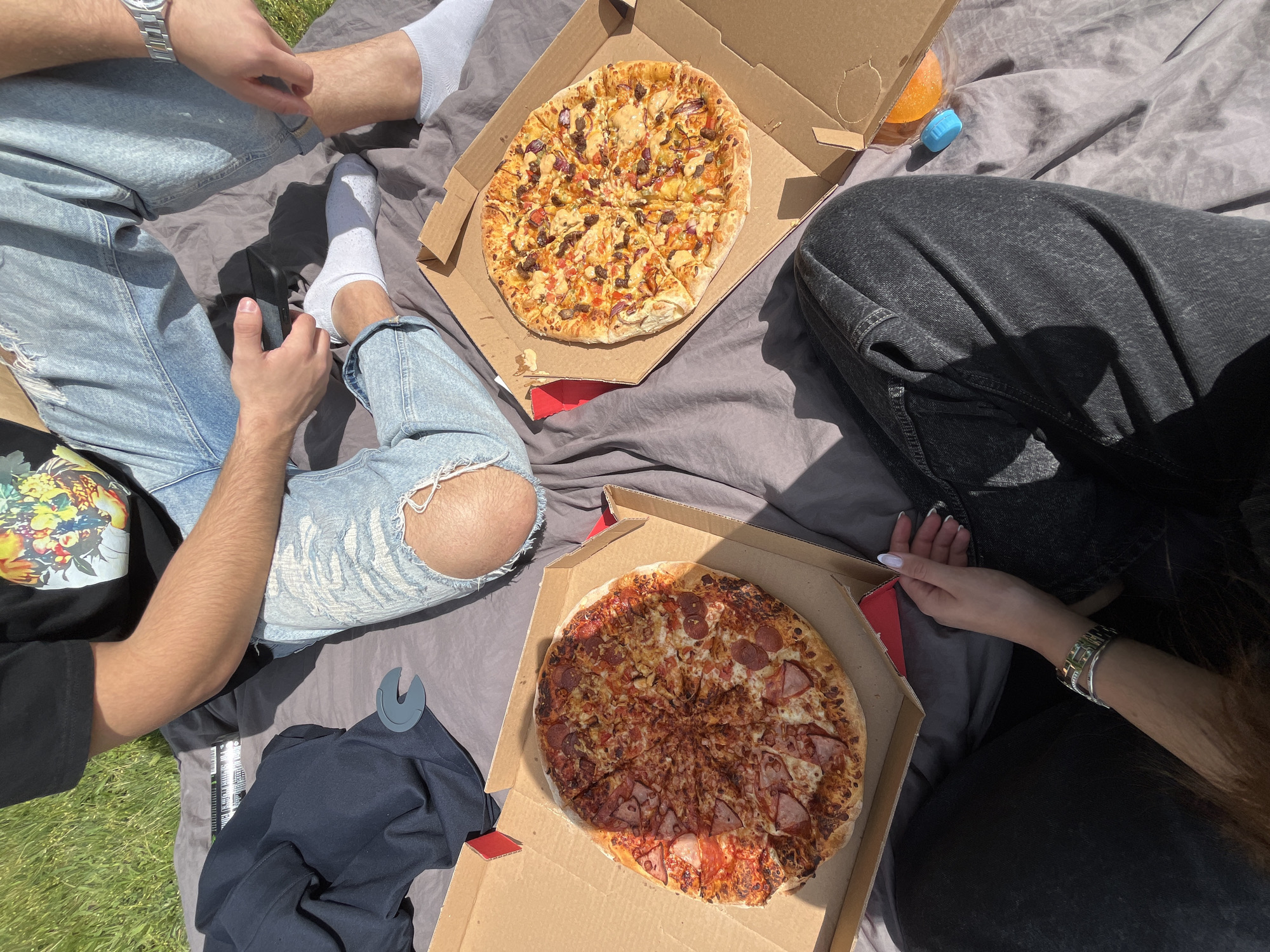 A high-angle shot depicts three people sitting on a grey blanket, seemingly enjoying a picnic with pizza. Two pizza boxes are open, each containing a different type of pizza. One pizza features toppings such as meat and onions, while the other is topped with pepperoni and ham. The people wear casual attire: one person is in ripped light blue jeans and white socks, another in dark grey jeans, and the third in light blue jeans and white socks with a watch. A dark-colored jacket is visible. The setting appears to be outdoors on grass, suggesting a park or green space. The exact location in Sofia, Bulgaria, is not evident from the visual information provided.