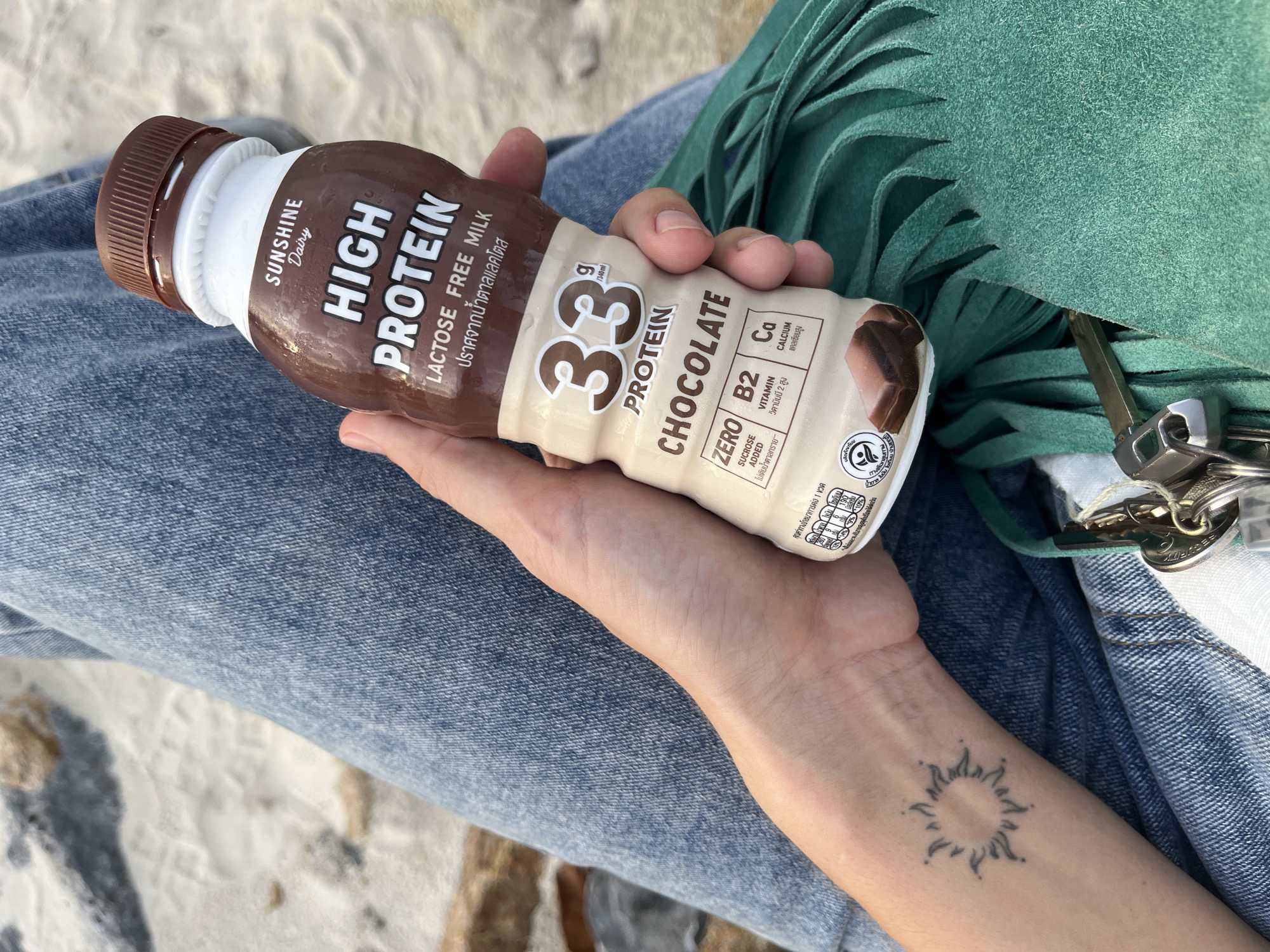 The image displays a close-up perspective of a person's hand holding a brown and white protein drink bottle, set against a sandy background in ตำบลเกาะพะงัน, Thailand. The person's right hand grips a bottle of "Sunshine Dairy HIGH PROTEIN LACTOSE FREE MILK Chocolate," which advertises "33 g PROTEIN / 340ml," "ZERO SUCROSE ADDED," "VITAMIN B2," and "CALCIUM (Ca)." Condensation is visible on the bottle's surface. On the person's left wrist, a minimalist tattoo depicting a sun with radiating lines is present. The person is wearing light blue denim jeans, partially visible on the left and lower right of the frame. Resting on the jeans and a teal/turquoise suede or faux suede bag with fringe detailing, a set of keys including a silver and black car key fob and other metallic keys on a keyring are visible. The background consists of light-colored sand with faint footprints and scattered small, dark stones or debris, indicative of a beach or outdoor sandy area.