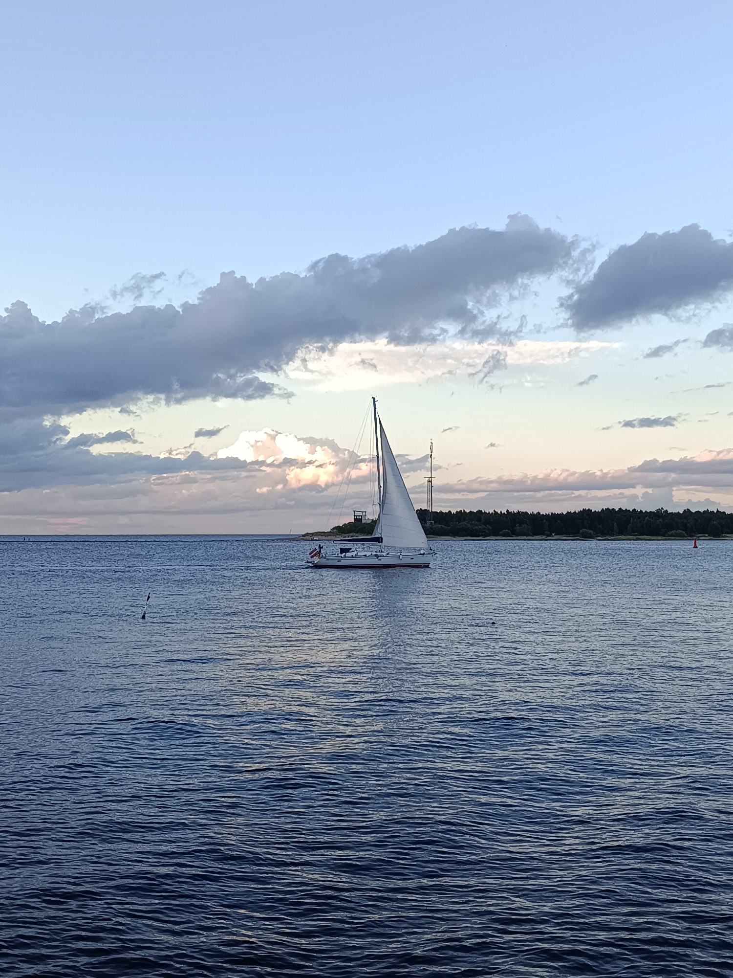 A solitary sailboat with white sails glides across the calm, dark blue water of the Daugava River in Riga, Latvia. The sky above is a soft gradient of blue, transitioning to pale yellow and pink near the horizon, where dramatic, dark gray clouds are punctuated by illuminated patches of white and pink, suggesting the sun is setting or has recently risen. A thin line of green trees marks the distant shore, and a few structures, including a tall antenna and a lighthouse-like building, are visible on the land. The water exhibits subtle ripples, reflecting the subdued light.