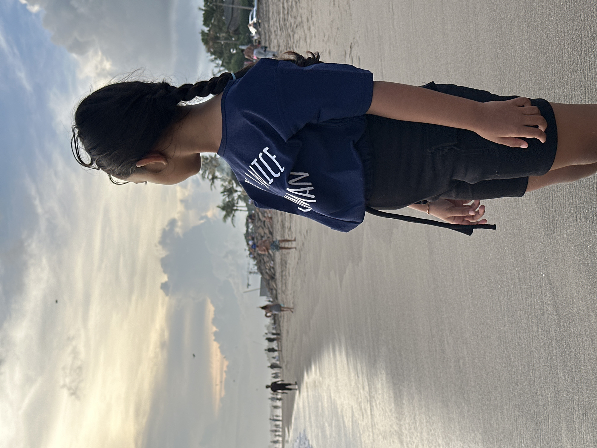 A young person stands on a sandy beach, likely in North Kuta, Indonesia, given the presence of palm trees and beachgoers in the background. The individual has dark hair styled in braids and is wearing a dark blue t-shirt with white lettering, and dark shorts. The person appears to be looking at the ocean. The background includes a beach with other people walking or standing near the water's edge. The sky is cloudy, with some areas appearing brighter, possibly indicating a sunset or sunrise.