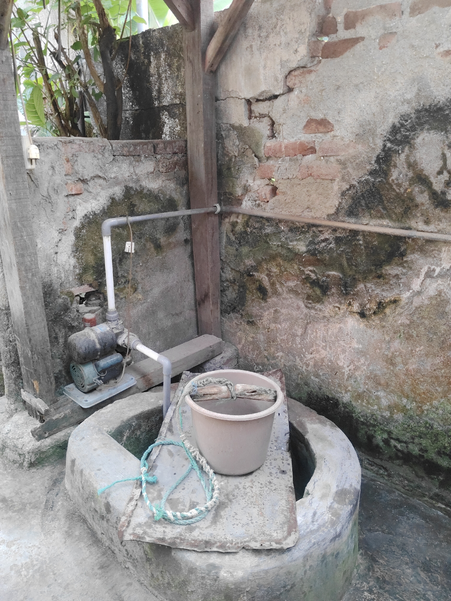 A well with a water pump and bucket is shown. The well is made of concrete and is partially covered by a wooden plank. A beige bucket with a rope and a piece of wood is placed on the plank. A water pump is connected to a pipe that runs along a brick wall. The wall is damaged and has moss and water stains. Trees and foliage are visible in the background. The scene appears to be outdoors in a humid environment, likely in a residential area of Surabaya, Indonesia, given the presence of a water well and pump, common in areas with inconsistent water supply.