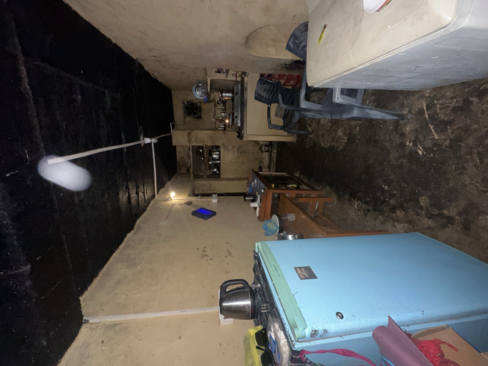 The image depicts a dimly lit, narrow room that appears to be a kitchen or living space within a residential dwelling in Haini, India. The walls are a rough, tan plaster, and the ceiling is a dark, textured material that appears to be tarred or heavily stained. The floor is uneven and appears to be made of packed earth or dirt, with visible debris and what looks like water or mud.
On the left side of the room, a light blue refrigerator, branded "FAIRLAND," is prominently positioned. On top of the refrigerator sits a silver electric kettle and some other objects. To the right of the refrigerator, a wooden table with a bench runs along the wall. On this table are several glasses, a plate, and a kettle.
Further into the room, a gas stove with burners is visible, set on a counter. Shelves above the stove hold various kitchen utensils and containers. Two black plastic chairs are placed in front of the stove area, and another chair is partially visible in the foreground on the right. A red gas cylinder is situated behind one of the chairs.
In the background, a diamond-shaped blue object is mounted on the wall, possibly an air freshener or decorative item. A single light bulb illuminates the space, casting shadows and highlighting the worn and somewhat cluttered nature of the room. The overall impression is one of a basic, functional living space in a rural or semi-urban setting.