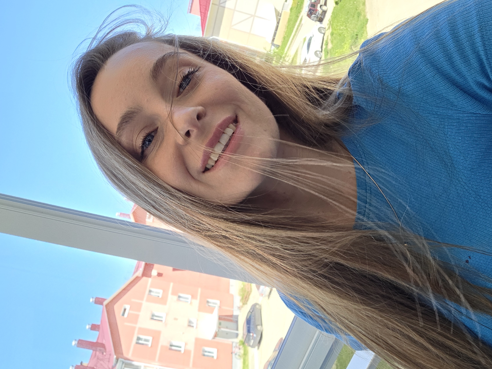 A young woman with long blonde hair is smiling and looking directly at the camera. She is wearing a blue top. Behind her, a window frames a view of a multi-story building with a red roof. The building has white-framed windows. The sky is bright blue. A car and a small tractor can be seen in the distance. This image was taken in Birobidzhan, Russia.