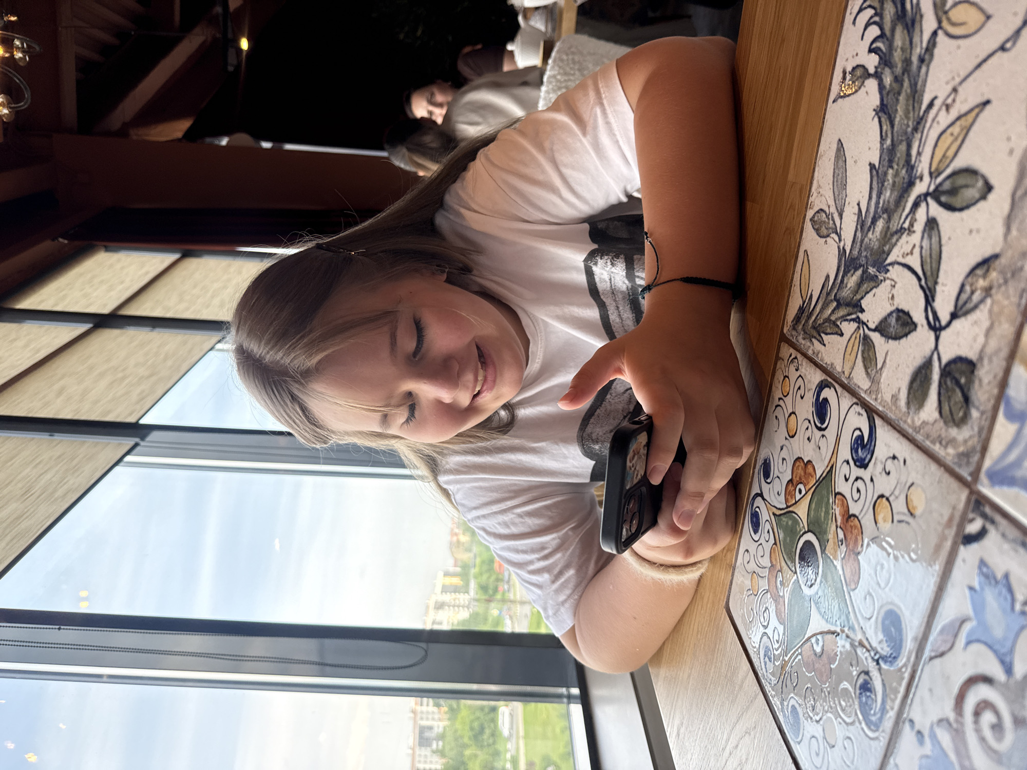 A young woman is seated at a table with decorative tiles in what appears to be a restaurant or cafe. She is looking at a smartphone she is holding in her hands. Outside the window, a cityscape is visible, identified as Veliky Novgorod, Russia based on contextual evidence. The restaurant setting includes other patrons visible in the background. The lighting is bright, suggesting it is daytime. The woman is wearing a white t-shirt and has long hair. She is wearing a bracelet. The table is both wood and tile, with the tile being multicolored with floral patterns.