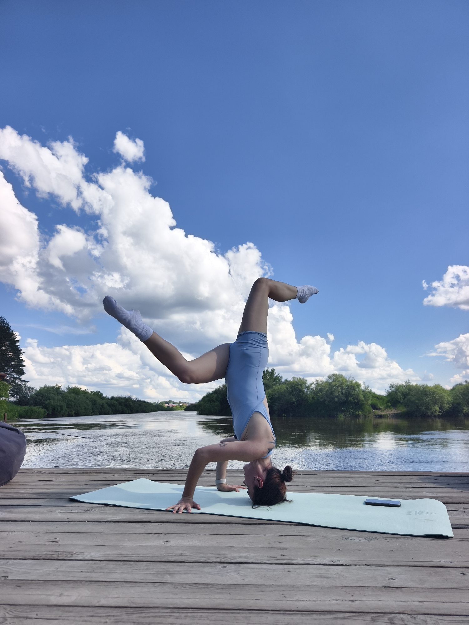 A person is performing a headstand yoga pose on a light blue yoga mat placed on a wooden dock. The person is wearing a light blue leotard and white socks. Their head is on the mat, supported by their hands, with their legs extended upwards. One leg is bent at the knee, and the other is straight. The person has dark hair styled in a bun. A dark gray beanbag chair is partially visible on the left side of the dock. A black smartphone is placed on the right side of the yoga mat. The dock is situated next to a body of water, likely a river, with a tree-lined shore on the opposite bank. The sky is blue with scattered white clouds. The location is identified as Onochino, Russia.