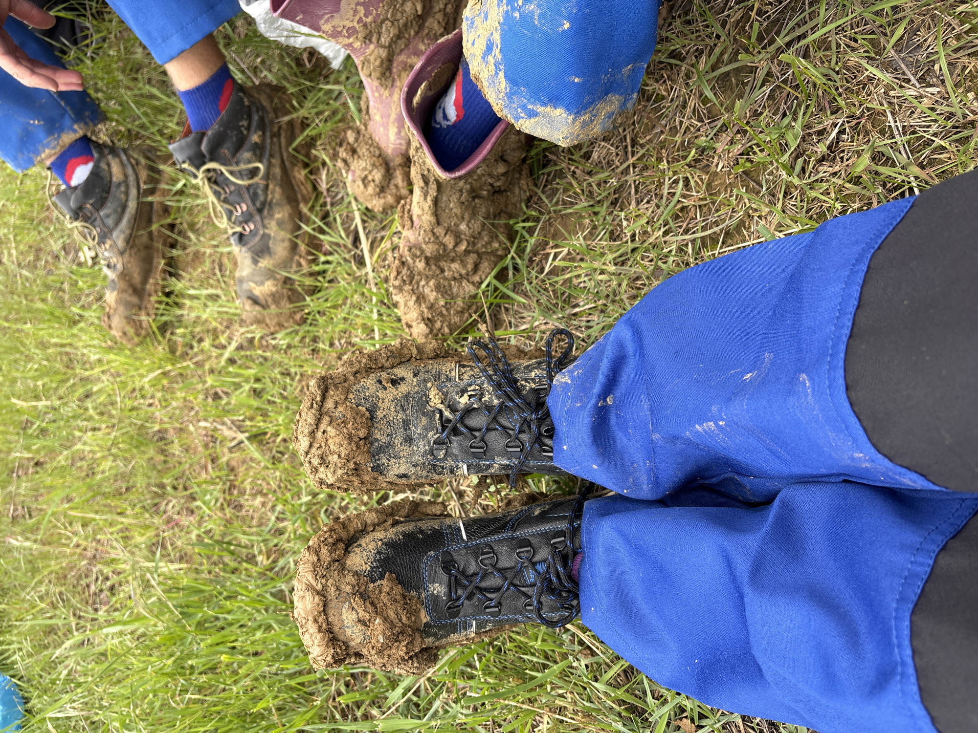 The image depicts a grassy area with several pairs of muddy boots. Three pairs of boots are visible, covered in dark brown mud. Two people are wearing blue work pants with black side panels. The other pair of boots is buried in mud, but the top part of a sock that is navy blue, red and white is visible. The location is most likely not Pushkino, Ukraine due to the current war and occupation.