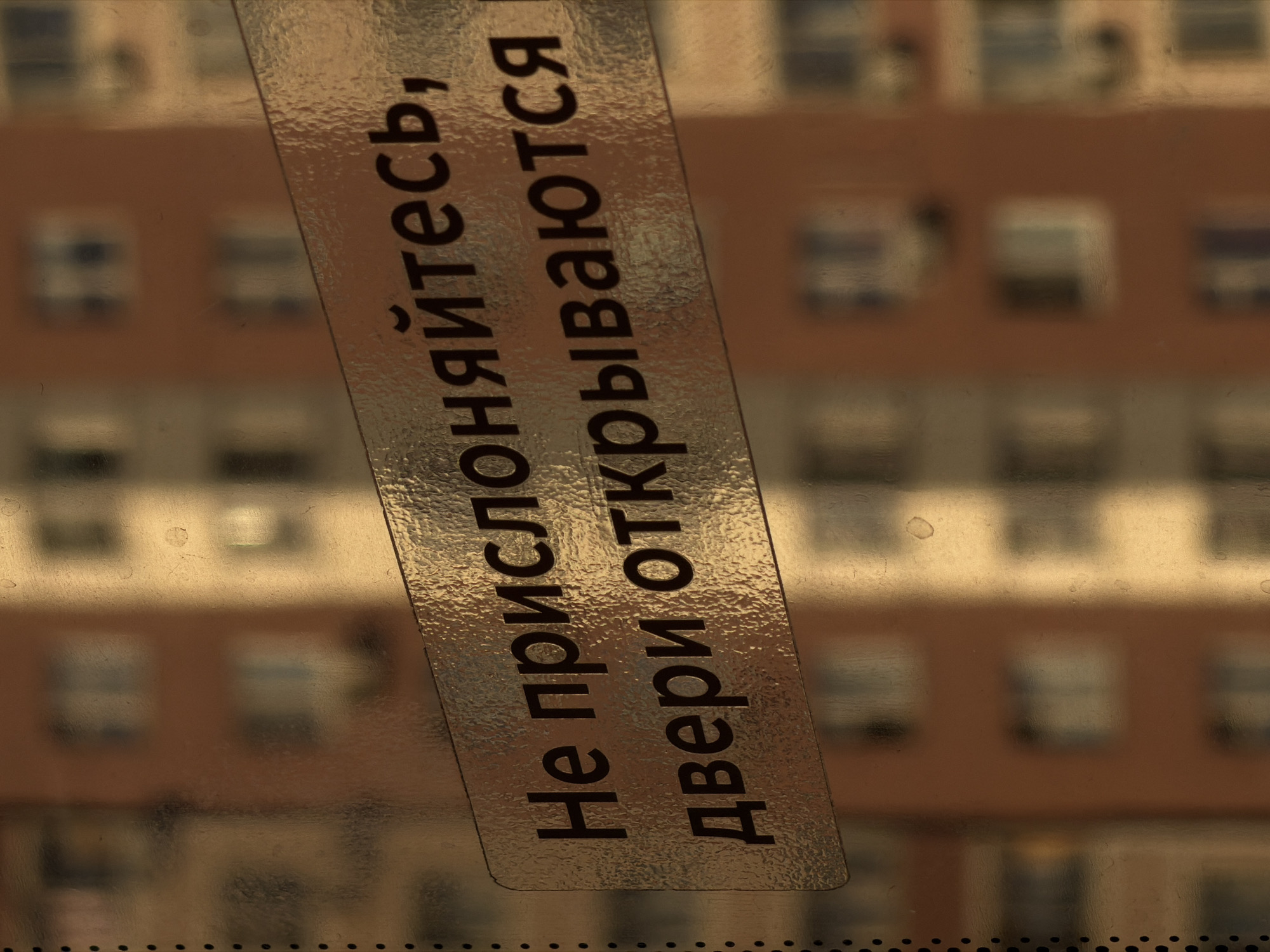 This media file captures a close-up view of a sign attached to a window or glass surface. The sign is made of a textured, metallic-colored material with black text. The background is a blurred image of a building, suggesting an urban or residential setting. The building appears to have multiple floors with rows of windows, rendered in muted tones of brown and beige, indicating it might be daytime but not necessarily sunny.
The sign itself is oriented diagonally, with the text running from the bottom left to the top right. The text is in Russian and reads: "Не прислоняйтесь, двери открываются" which translates to "Do not lean, doors open." This is a functional warning sign, likely placed on a glass door or partition to prevent accidents. The textured surface of the sign and the glass it's on create a slightly distorted and diffused effect on the background image.
The overall impression is that of an everyday scene, possibly in an apartment building or a public space with glass doors. There are no people visible, and the focus is entirely on the sign and its immediate surroundings. The lighting appears to be ambient, and there are no strong shadows or highlights that would indicate a specific time of day beyond it being light. The location context of Butovo, Russia, helps to identify the specific cultural and geographical setting of this common warning.