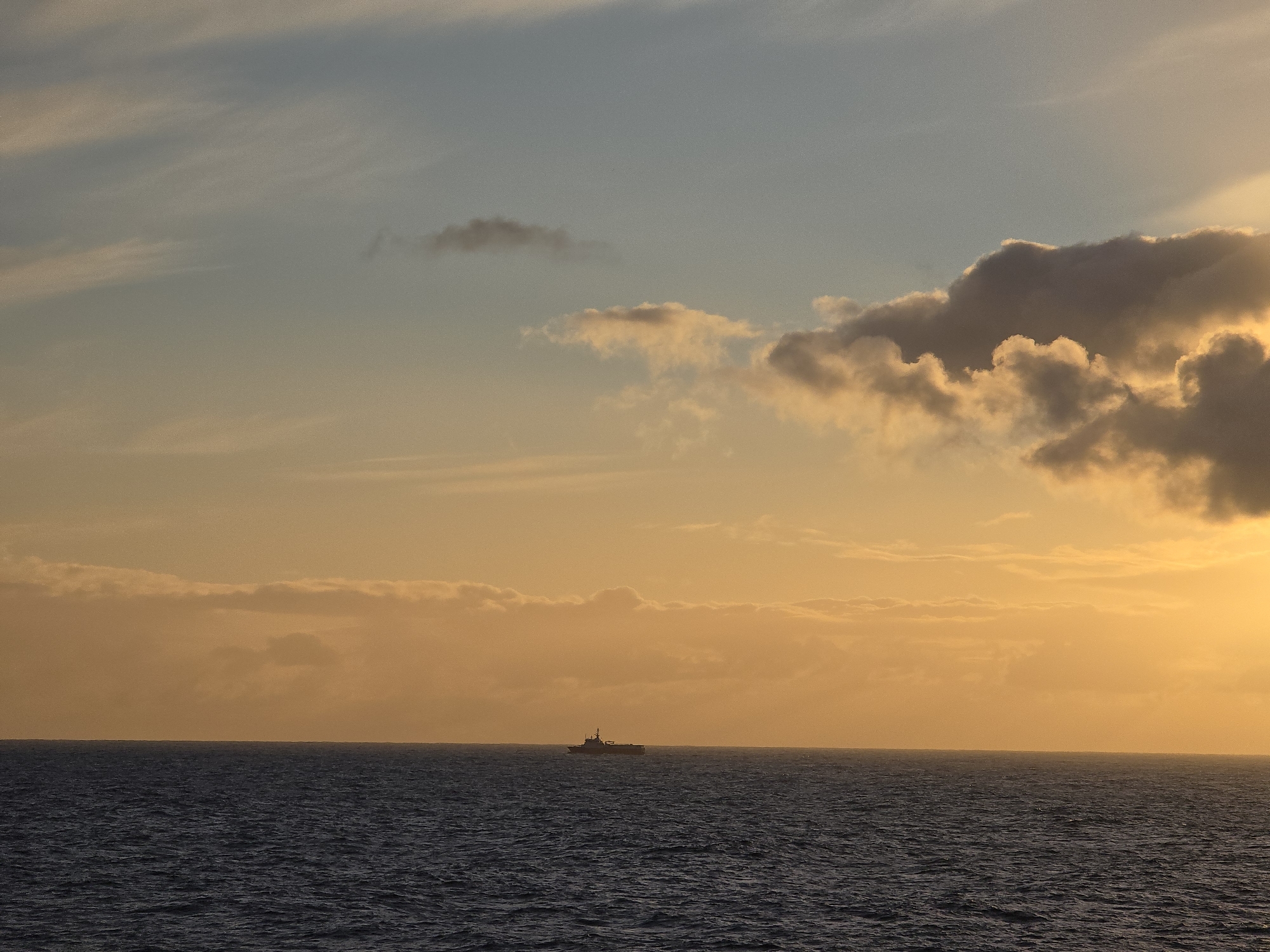The image depicts a body of water with small waves under a sky with clouds tinted with hues of orange and yellow, suggesting a sunrise or sunset. A silhouette of a ship is visible on the horizon. The location is Unknown, Unknown.