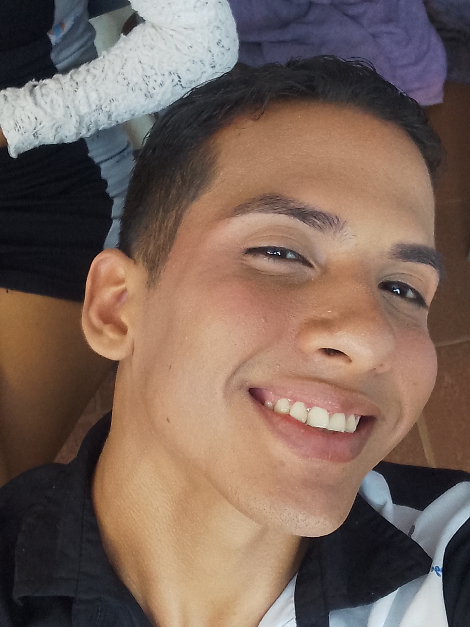 A head-and-shoulder close-up photograph captures a young adult male with a medium-dark complexion, smiling broadly. His dark, short-cropped hair frames his face, with a receding hairline visible on the forehead. His dark eyes, accentuated with visible eyeliner on both upper and lower lids, are directed slightly upward and to the viewer's left. His nose is of moderate width with a rounded tip. A wide smile reveals an upper and lower set of white, healthy-looking teeth. The subject is wearing a black collared shirt, with a distinct white and grey striped pattern visible on the lower right side of the garment. To the upper left of the frame, a white long-sleeved garment with a detailed lace or crochet pattern is partially visible, likely worn by another person. Below this, a segment of dark fabric is present. In the upper right background, a blurred purple fabric is visible, while the lower right displays a reddish-brown, textured surface, possibly a floor or wall. The lighting appears even, illuminating the subject's face directly. No specific geographical indicators are present in the image to confirm a location such as Porlamar, Venezuela.