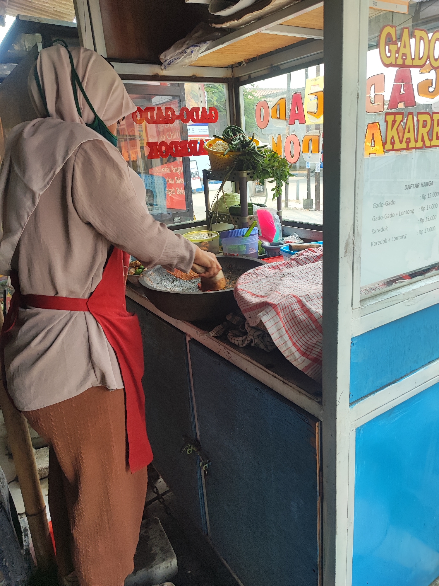 The image depicts an outdoor scene in Bekasi, Indonesia, showing a street food vendor preparing food. The vendor, a woman wearing a hijab and a red apron over a light-colored tunic, is seen from the side, her back partially to the camera, actively engaged in cooking in a large wok-like pan. She is wearing a light brown hijab, a beige long-sleeved top, a red apron tied around her waist, and loose brown pants. A dark green face mask is visible. The vendor is standing behind a food cart, which has signage indicating menu items like "GADO-GADO" and "KAREDAK." A price list, labeled "DAFTAR HARGA," is affixed to the cart. The cart is constructed of blue and white painted metal and wood. On a shelf within the cart, there are various ingredients and cooking utensils, including a basket of fresh green vegetables, plastic containers, and a patterned cloth covering some items. The background suggests an urban environment with some buildings and possibly other street activities. The overall impression is a snapshot of daily commerce in a bustling Indonesian city.