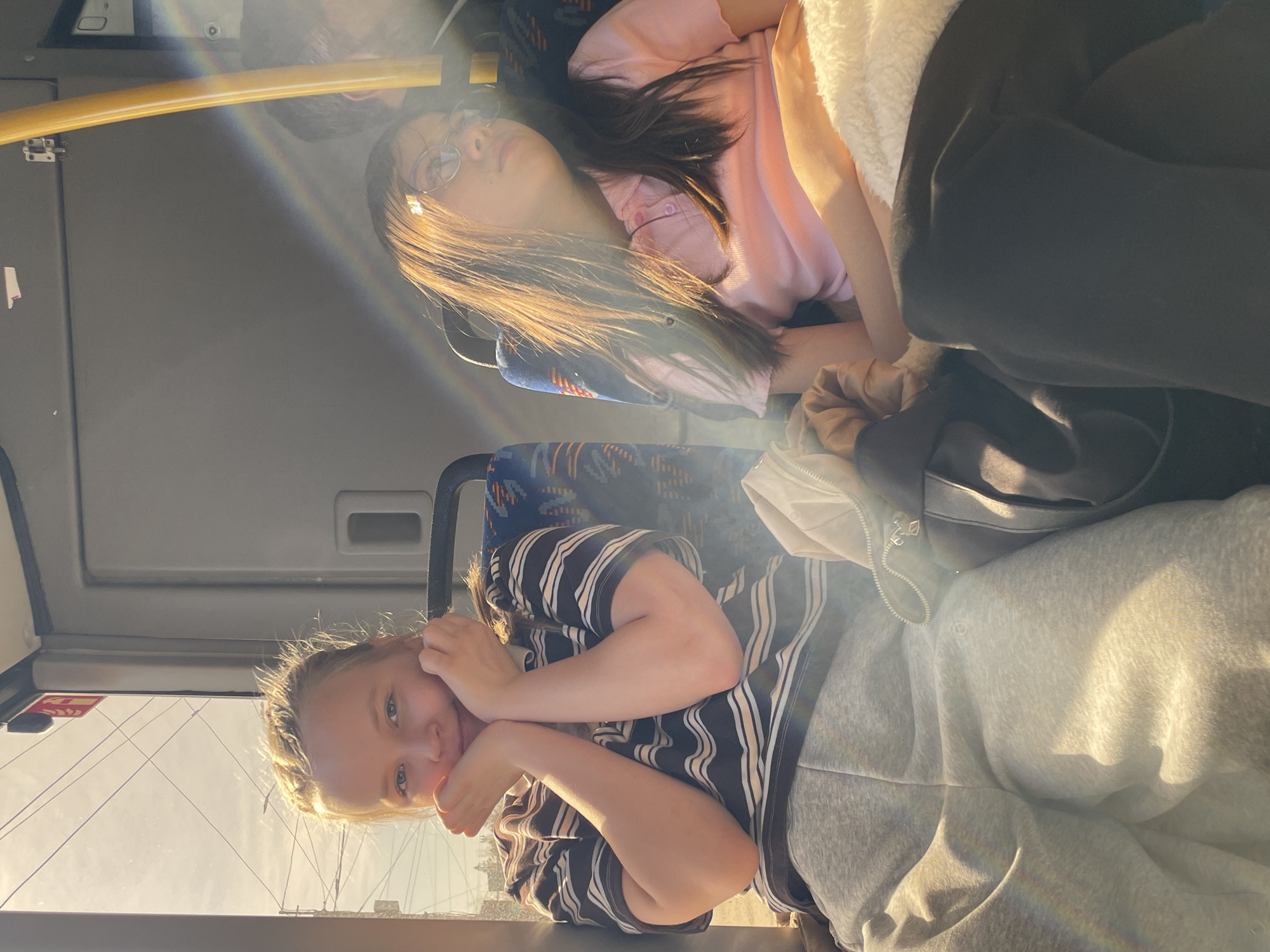 Two girls are sitting on a bus. The younger girl, in the foreground, wears a striped shirt and grey sweatpants. She rests her face on her hands and looks at the camera. Behind her, the other girl is asleep on the seat. She wears glasses and a pink shirt. The bus appears to be in Izhevsk, Russia. There is a rainbow effect in the upper left corner of the photo.