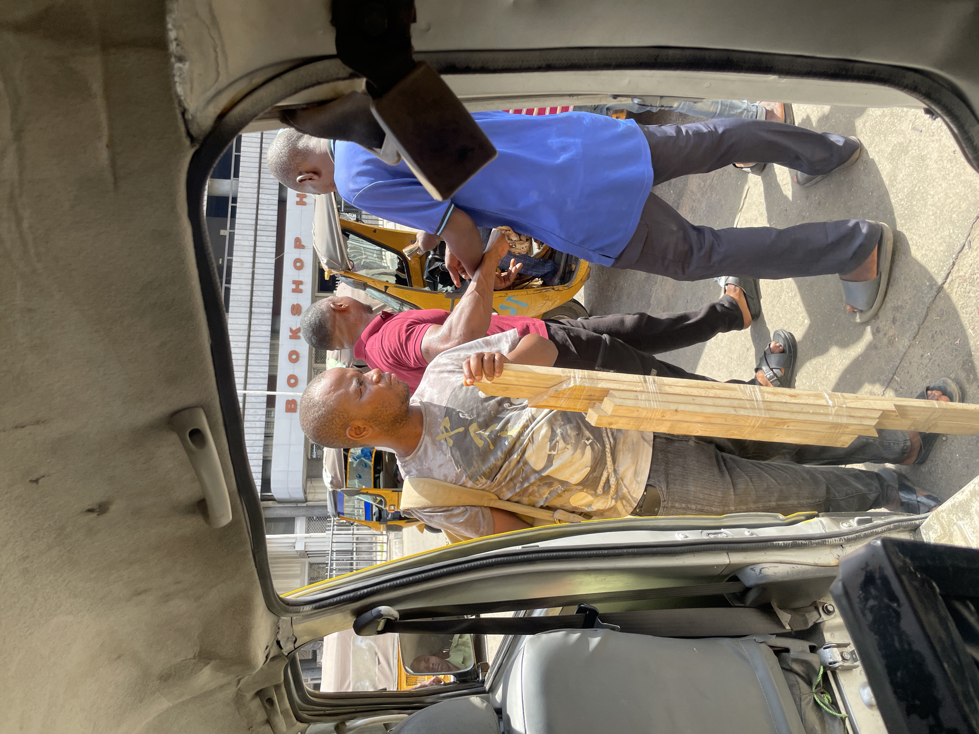 The image, taken from inside a vehicle looking out, captures a sunny urban street scene in Lagos, Nigeria. The bright daylight creates strong shadows on the ground.
In the foreground, a man with short hair and a beard, wearing a light-colored t-shirt with a graphic design and dark pants, is carrying a bundle of several light-colored wooden planks, wrapped in clear plastic. A light-colored strap from a bag or satchel is visible across his chest.
Behind him, another man in a red t-shirt and dark pants stands. Further back, a third man wearing a blue polo shirt and dark pants, with sandals on his feet, is visible. These men are positioned near what appears to be a yellow auto-rickshaw, a common sight in Lagos.
In the background, a building features a partial sign with vertical lettering that clearly reads "BOOKSHOP" at the bottom, though the top portion is obscured. The interior of the vehicle from which the photo was taken is partially visible, showing the headliner, a grab handle, and a rearview mirror. The scene depicts a casual street interaction or movement of goods in a busy city environment.