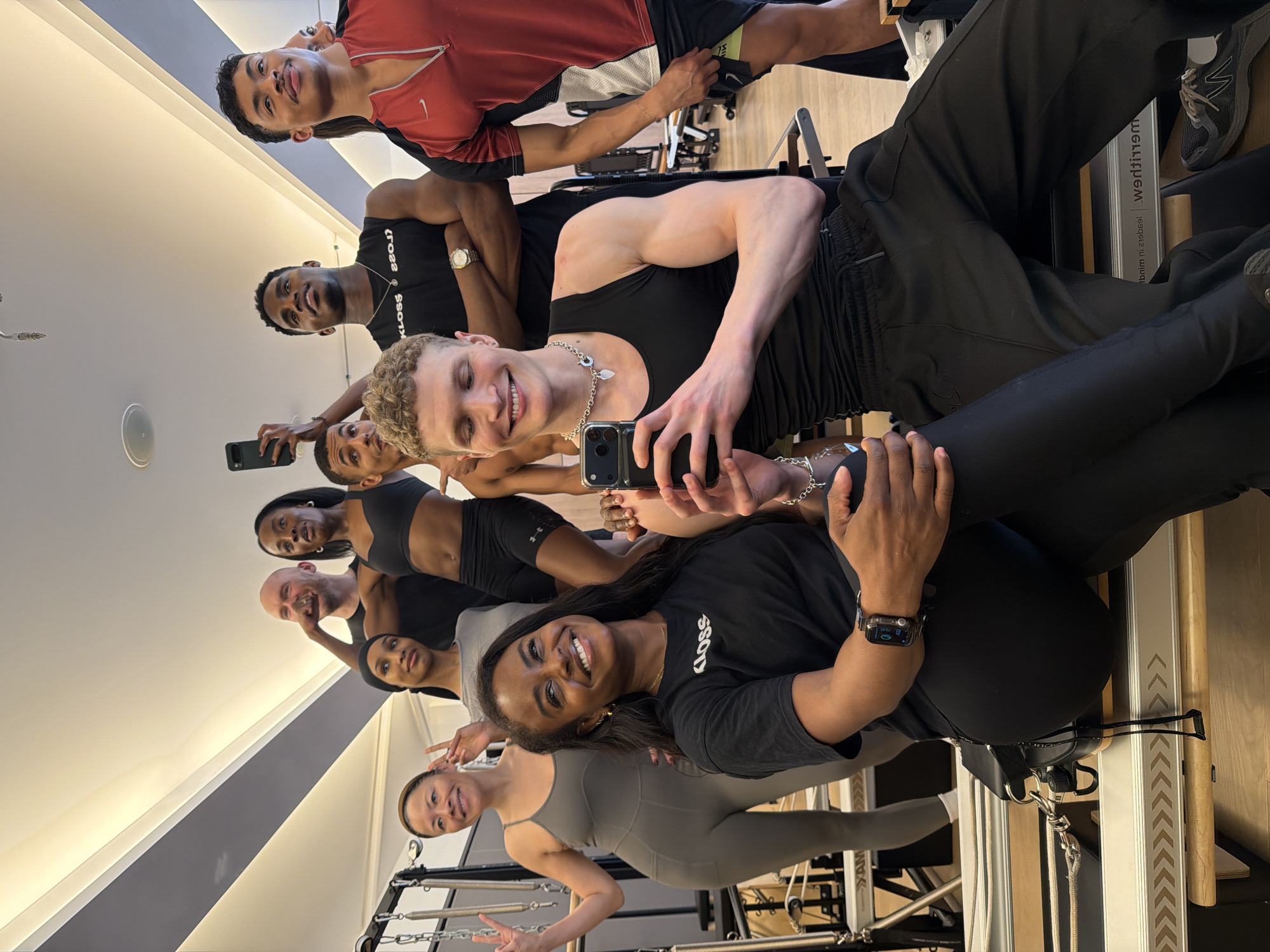 A group of nine people are posing for a selfie in a Pilates studio. The group is arranged in a line, with the person holding the camera positioned near the center. Most are smiling and looking at the camera. The Pilates studio has light-colored walls, light wood flooring, and the equipment is in clear view, including Pilates reformers. One person is wearing a red shirt with a Nike logo, and others are wearing black workout attire. A woman at the front is wearing a gray workout outfit and making a peace sign. A woman in a black t-shirt is also wearing a smartwatch. The t-shirt has the word "KLOSS" printed on it. The location is likely in London, United Kingdom.