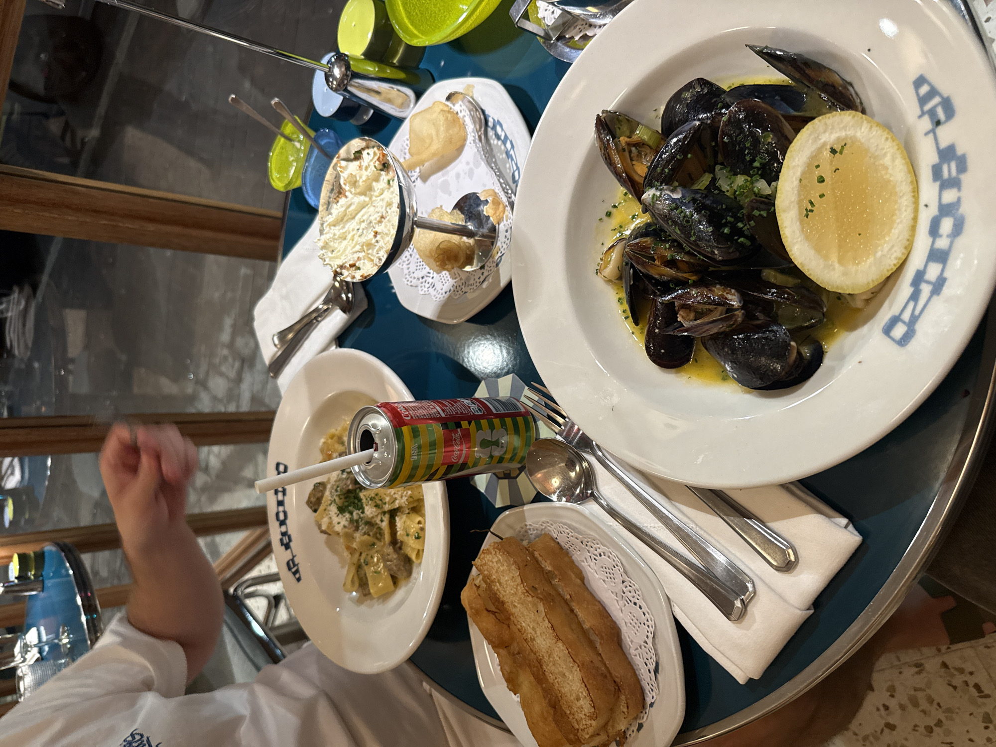 An overhead shot captures a dining scene at a table, likely in a restaurant or cafe in Cape Town, South Africa. The table, a reflective teal surface, is set with several dishes and drinks.
The main dishes include a large white bowl of steamed mussels in a light, herbed broth, garnished with a lemon wedge. Adjacent to it is a plate holding a pasta dish, possibly fettuccine or tagliatelle, coated in a creamy sauce with mushrooms, topped with grated cheese and fresh herbs. A colorful "Coke Studio" branded Coca-Cola can, featuring a vibrant red, yellow, and green pattern, is placed near the pasta, with a white straw inserted.
Smaller side dishes are also present: a small white rectangular plate holds several slices of toasted bread, resting on a lace doily. Another small plate features a creamy white dip or spread, garnished with herbs, served in a stylized metal cup, accompanied by two small fried dough pieces or savory pastries. Cutlery, including forks and spoons, are neatly placed on white napkins. In the background, two modern condiment dispensers with colorful (yellow and blue) bases are visible.
A person's arm and clenched fist, wearing a white shirt, are visible on the left side of the frame, suggesting an active diner. A reflection of a person's head and upper body can be seen in a dark, possibly mirrored or glass, surface in the upper left. The floor visible in the bottom right corner features a light-colored, patterned tile design. The plates have a stylized blue graphic on their rims.
The scene depicts a casual yet well-presented meal, possibly lunch or dinner, highlighting the food and beverages on offer.