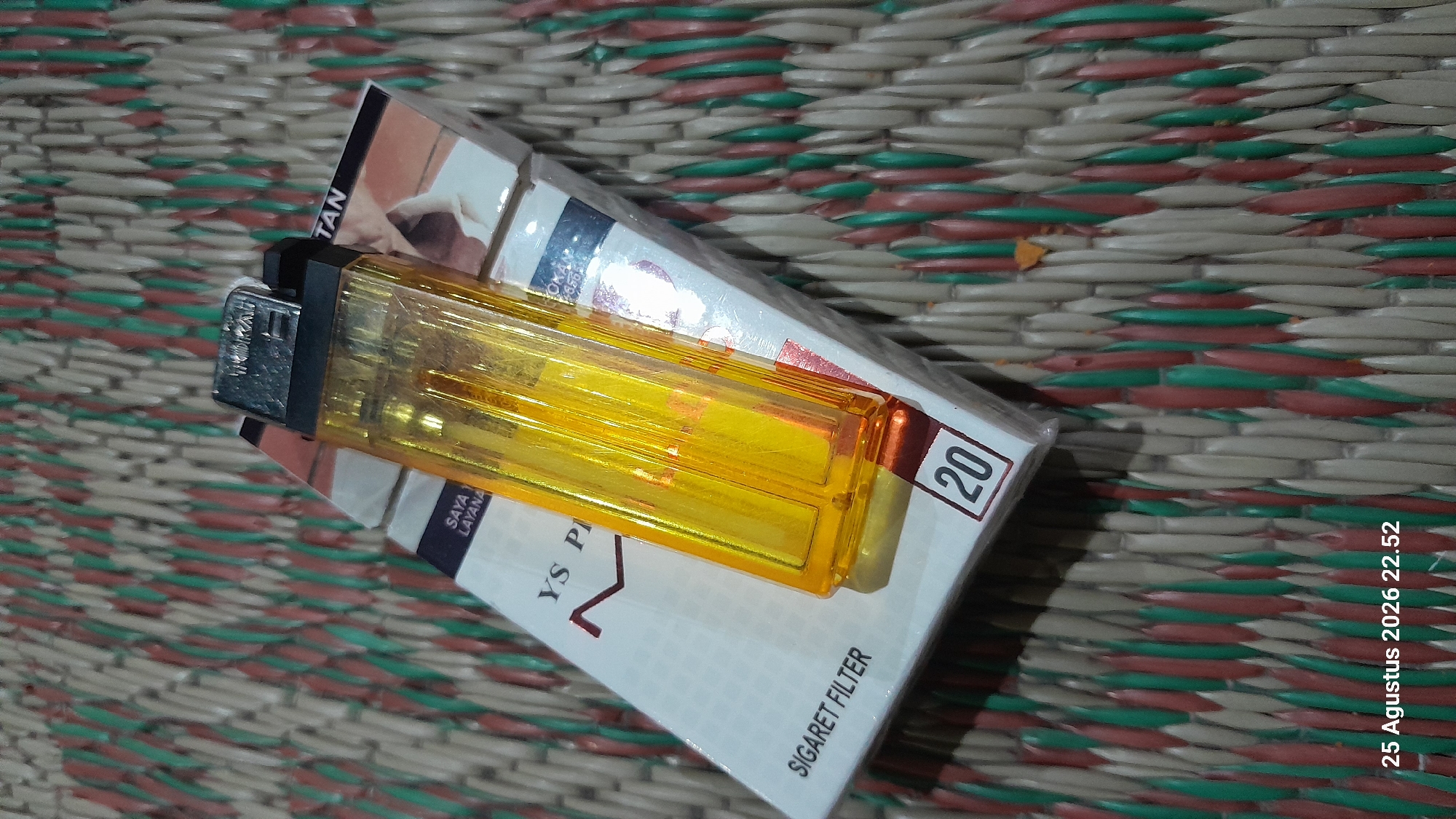 A yellow plastic lighter rests on a pack of cigarettes. The cigarettes are white with red and brown text and graphics. The pack has a label that reads "SIGARET FILTER" and "YS PR." The lighter is placed diagonally across the cigarette pack, with the top of the lighter near the top right corner of the pack and the bottom of the lighter near the bottom center. The background is a woven mat with alternating colored strands of red, green, and off-white. The timestamp "25 Agustus 2026 22.52" is visible in the bottom left corner of the image. The image appears to be taken indoors, likely on a table or floor. There are no people visible in the image.