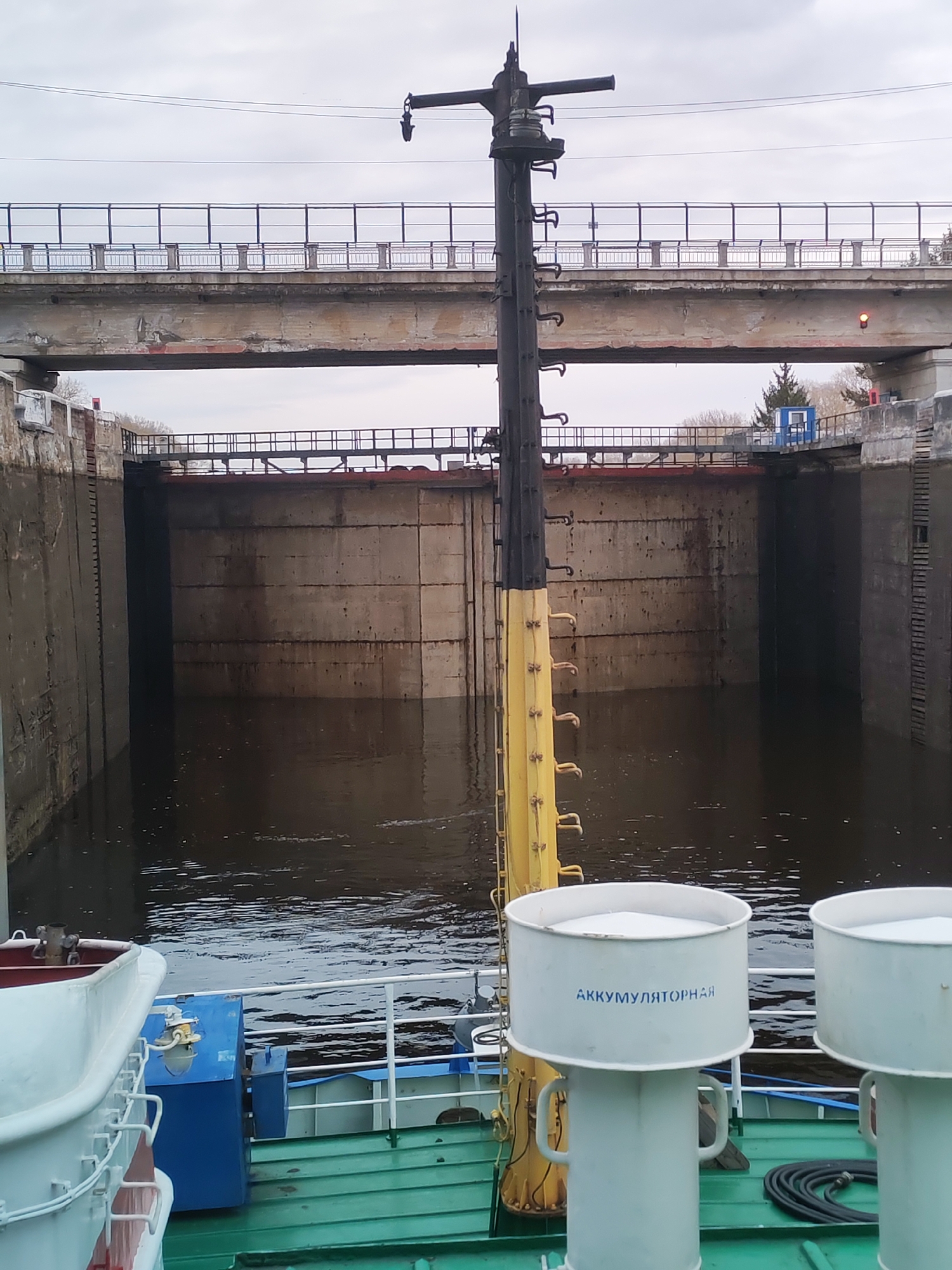 A medium, eye-level shot depicts a waterway lock, seen from the deck of a vessel with green-painted wood, with blue and white railings. Two white cylindrical structures, labeled "АККУМУЛЯТОРНАЯ", are in the foreground. A tall, black and yellow pole ascends from the vessel. In the background, the lock is constructed of concrete and holds dark water. Two bridges cross the lock, with overcast skies above. Based on the presence of Russian text and the provided location information, this is the lock structure in the city of Kopylovo, Russia.