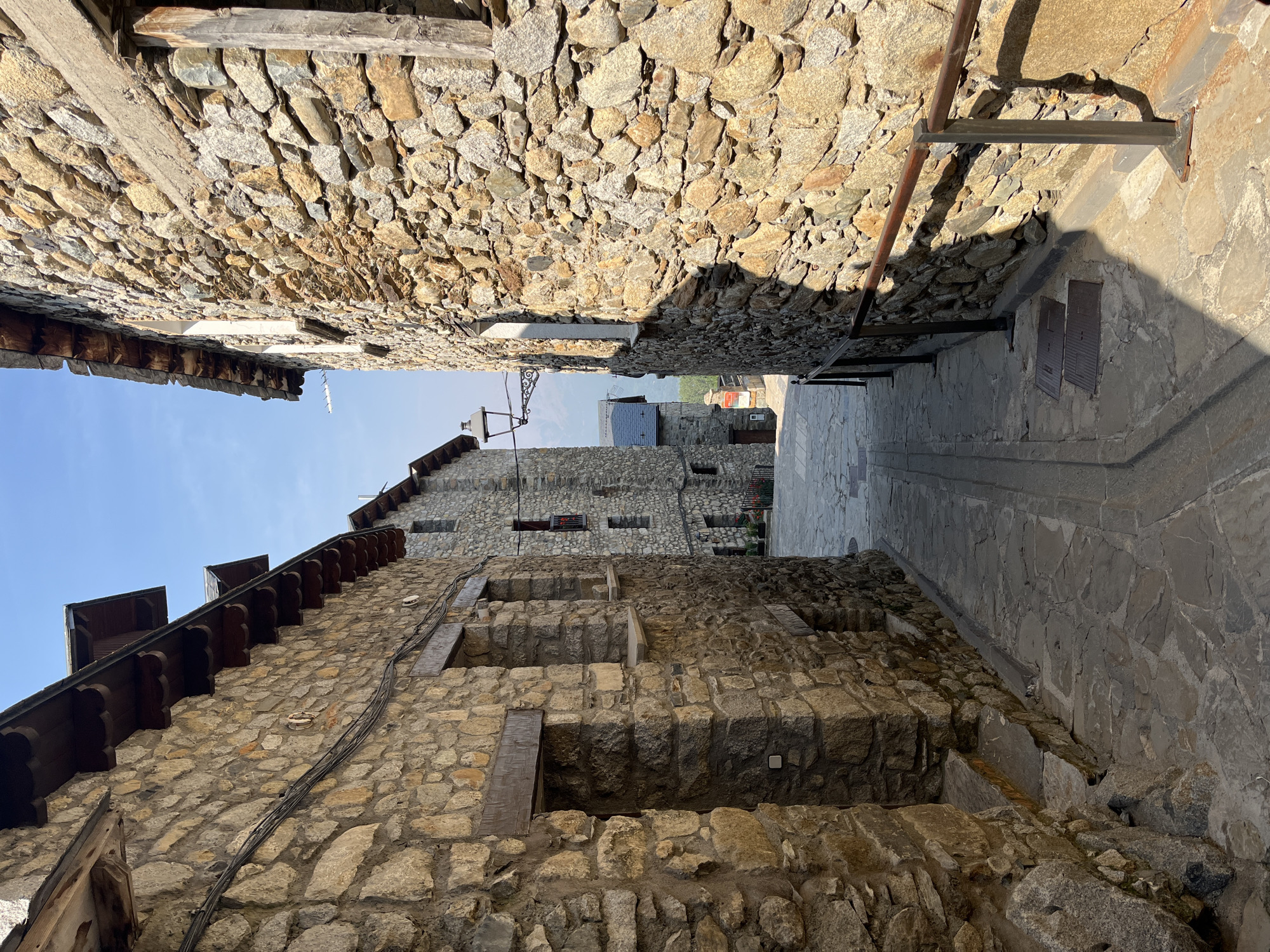 The image depicts a narrow, stone-paved alleyway in the historic center of Benasque, Spain. The buildings lining the alley are constructed of rough-hewn stone, with wooden eaves and window frames. The architecture suggests a traditional mountain village. The alley slopes gently upwards, leading the eye towards more stone buildings and a glimpse of a distant mountain landscape under a clear blue sky. Sunlight casts distinct shadows across the cobblestones and the stone walls, highlighting the texture of the materials. There are no people or animals visible in the scene. The overall impression is one of quiet antiquity and rustic charm.