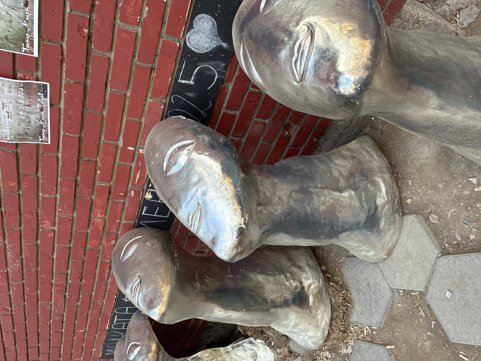 The image depicts a street scene in Plovdiv, Bulgaria, featuring a sculptural installation of several metallic, human-like heads. These heads are positioned on the ground against a brick wall that displays hand-written text and a price in Bulgarian currency. The ground surrounding the sculptures is paved with hexagonal tiles and scattered with debris. The overall impression is that of a public art piece integrated into the urban environment.